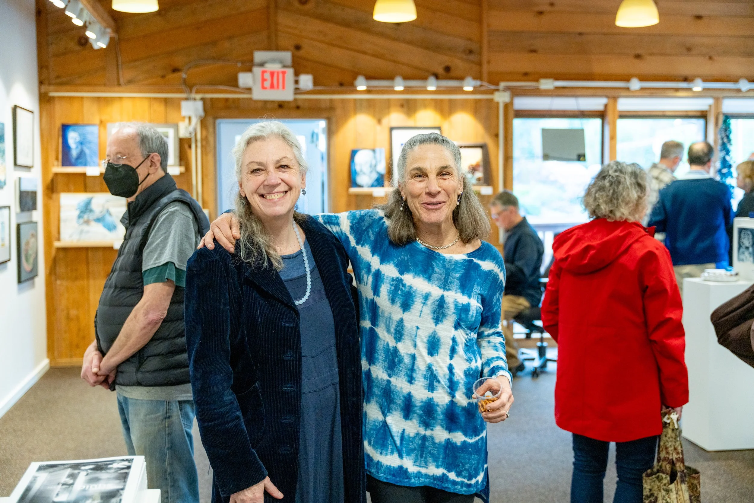 Two older women standing close together smiling at an art gallery, with other people browsing art in the background. One woman is wearing a blue tie-dye shirt and holding a drink, the other is wearing a navy jacket and a pearl necklace.