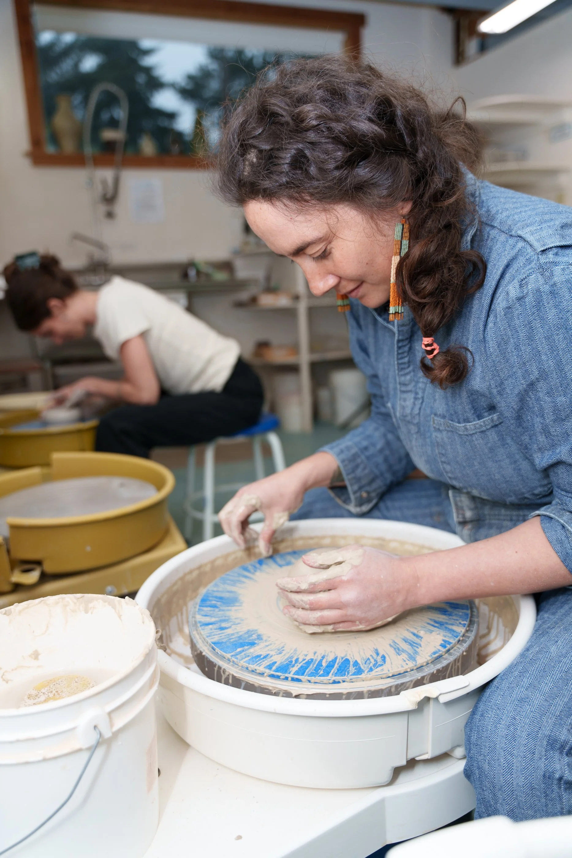 A woman shaping clay on a pottery wheel in a pottery studio, with another person working at a table in the background.