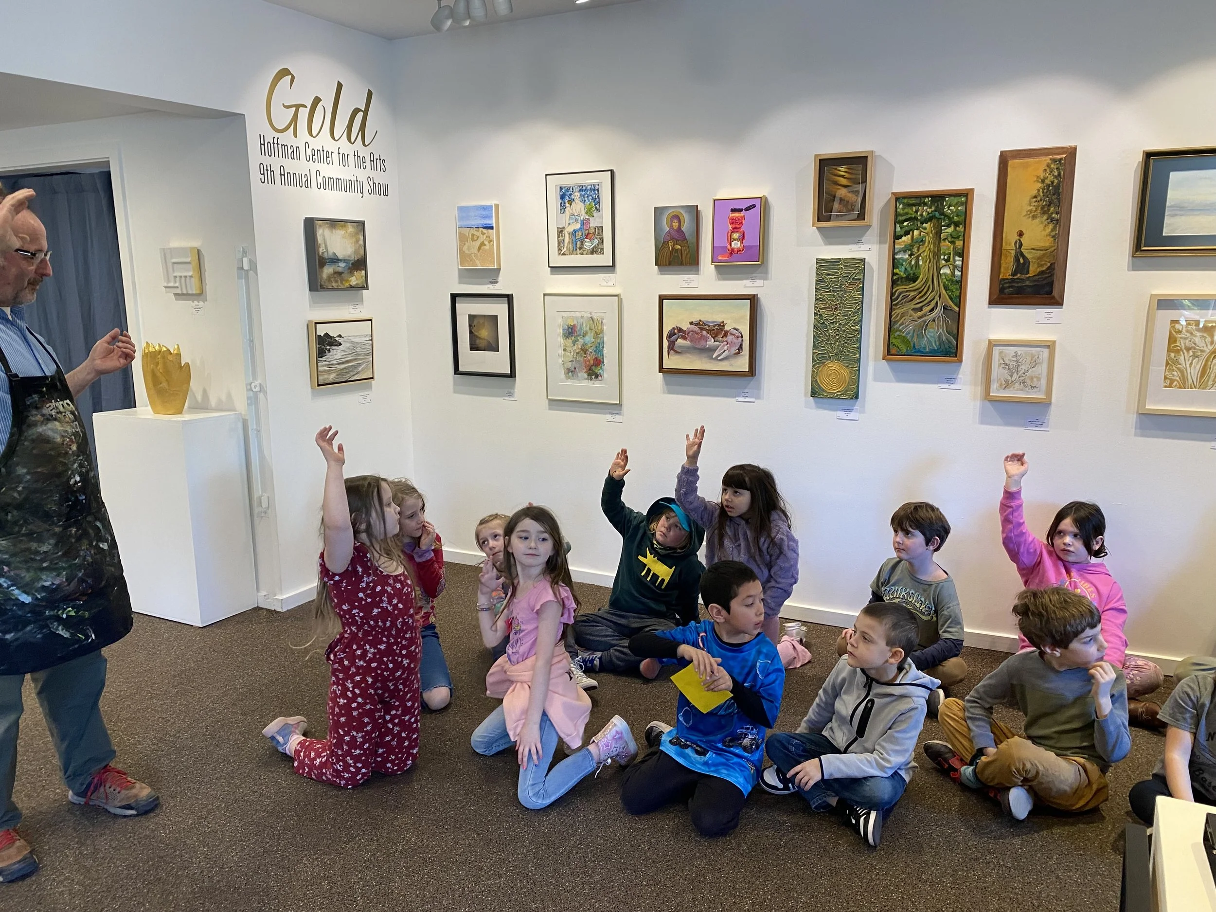 A group of children sitting on the floor and raising their hands during an art event at the Hoffman Center for the Arts, with artwork displayed on the white wall behind them.