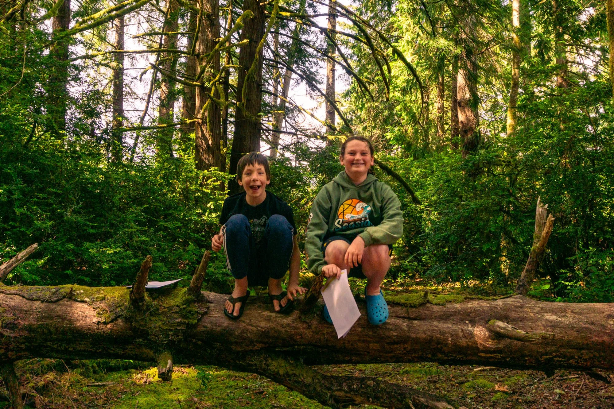 Two children, a boy and a girl, are sitting on a fallen log in a dense, green forest with tall trees and sunlight filtering through the leaves. The boy is smiling broadly, wearing a black t-shirt and dark pants, while the girl is smiling, wearing a green hoodie and Crocs, holding papers.