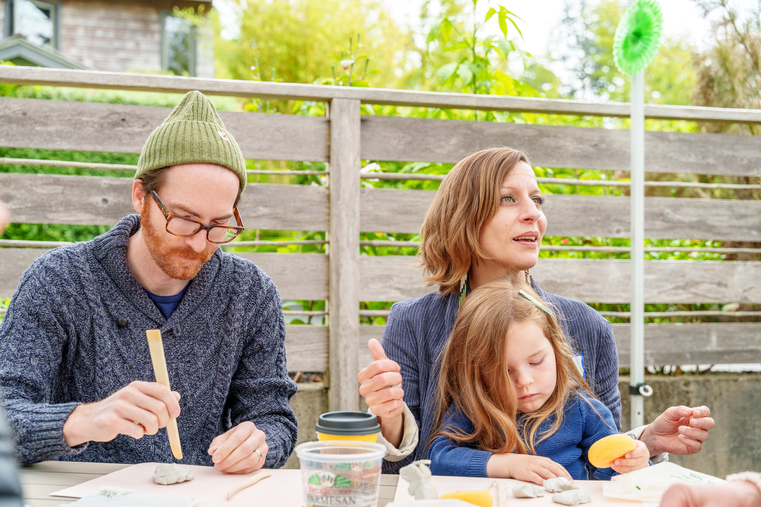 Family sitting outdoors at a table, making pottery with clay. A man with glasses and a green knit hat and a woman with shoulder-length hair are focused on their work, while a young girl with long hair is looking at a piece of clay. The background features a wooden fence and green foliage, with a colorful pinwheel on the side.