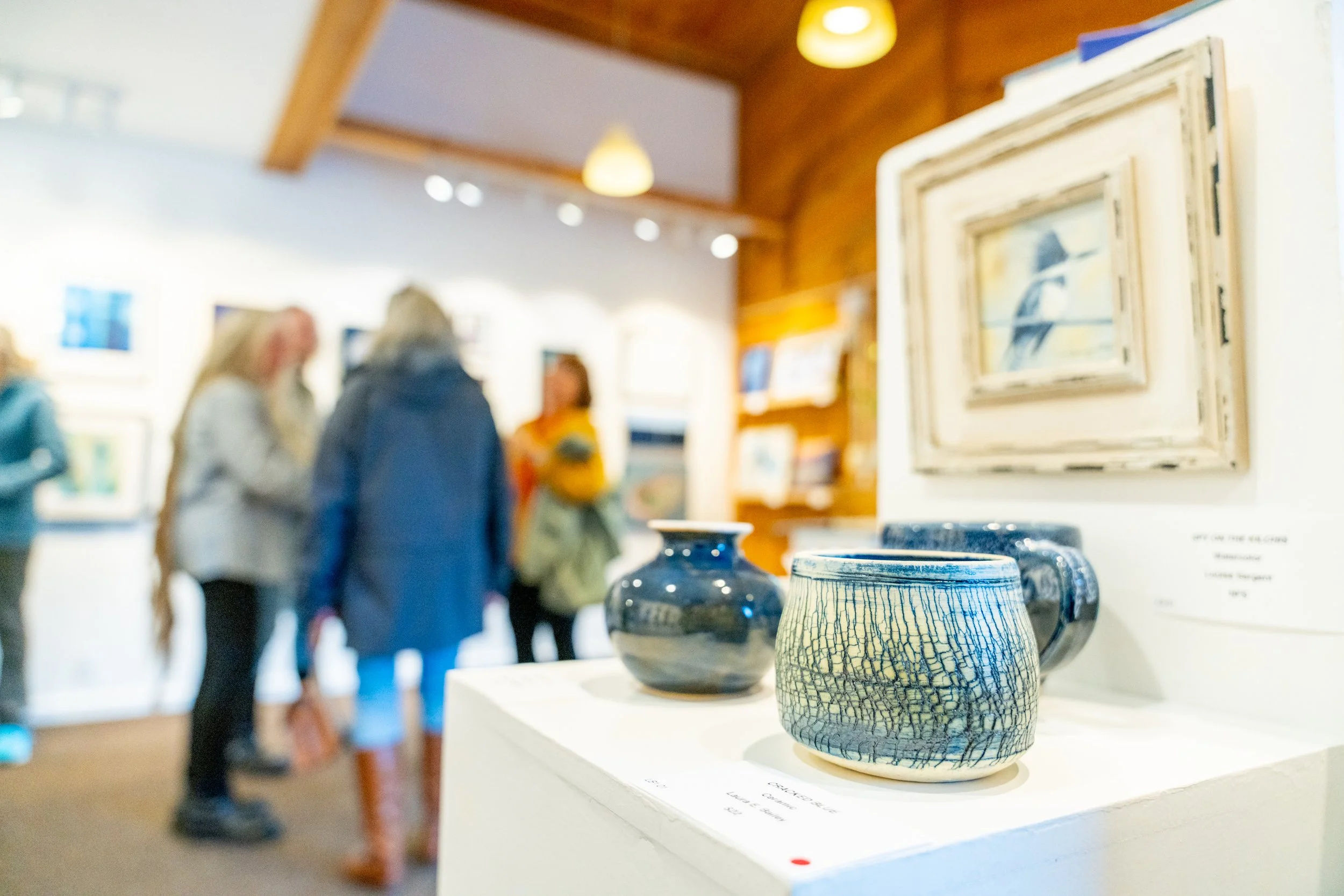 Ceramic vases displayed on a white pedestal at an art gallery with blurred visitors in the background.