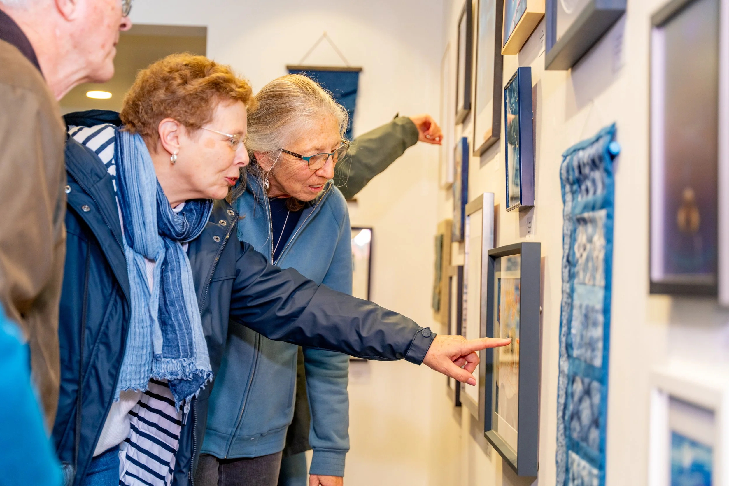 Four women closely observe and discuss artwork displayed on a gallery wall.