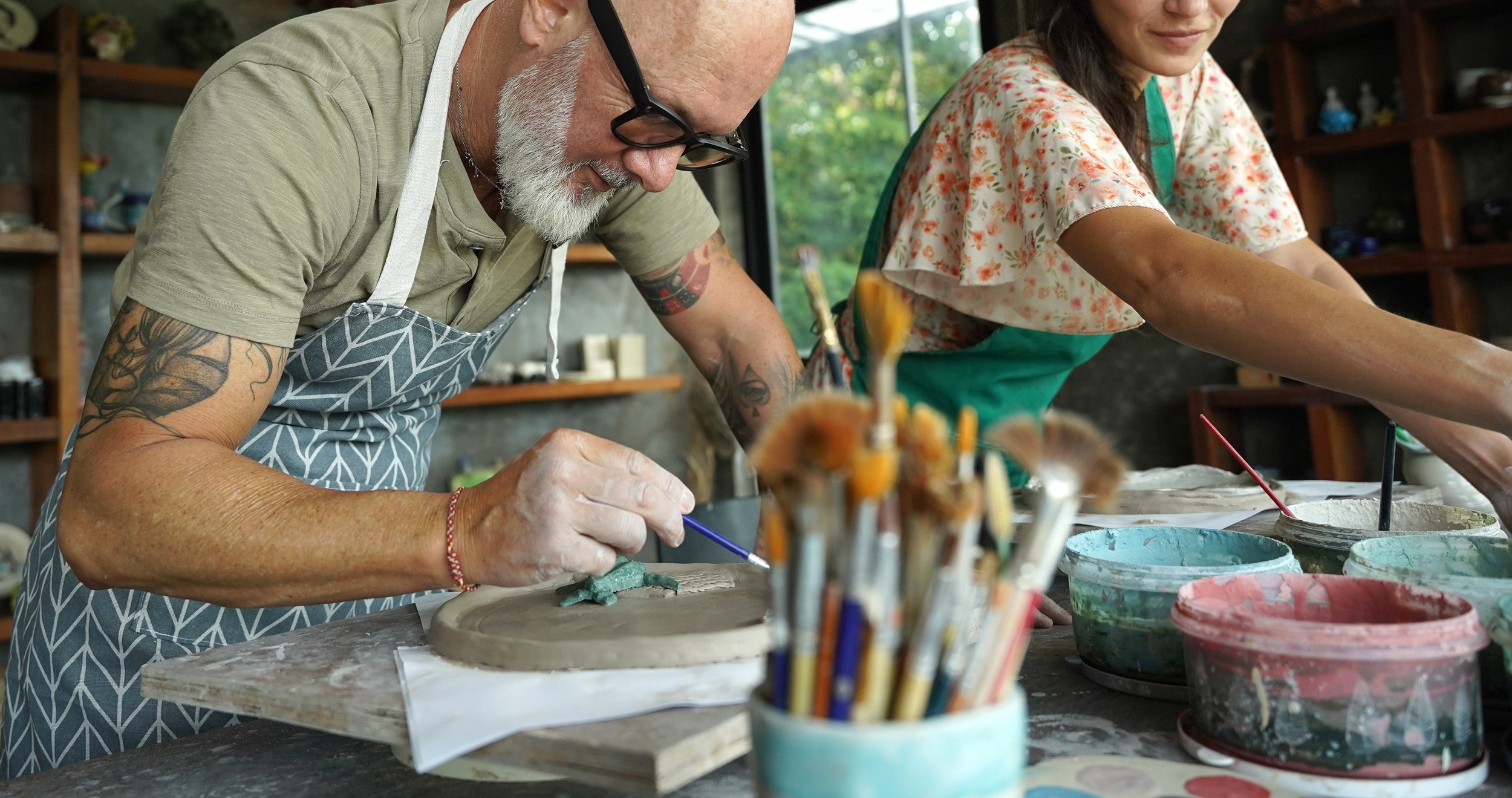 Two people working on pottery projects at a pottery studio, surrounded by bowls of paint and tools, with shelves of pottery pieces in the background.