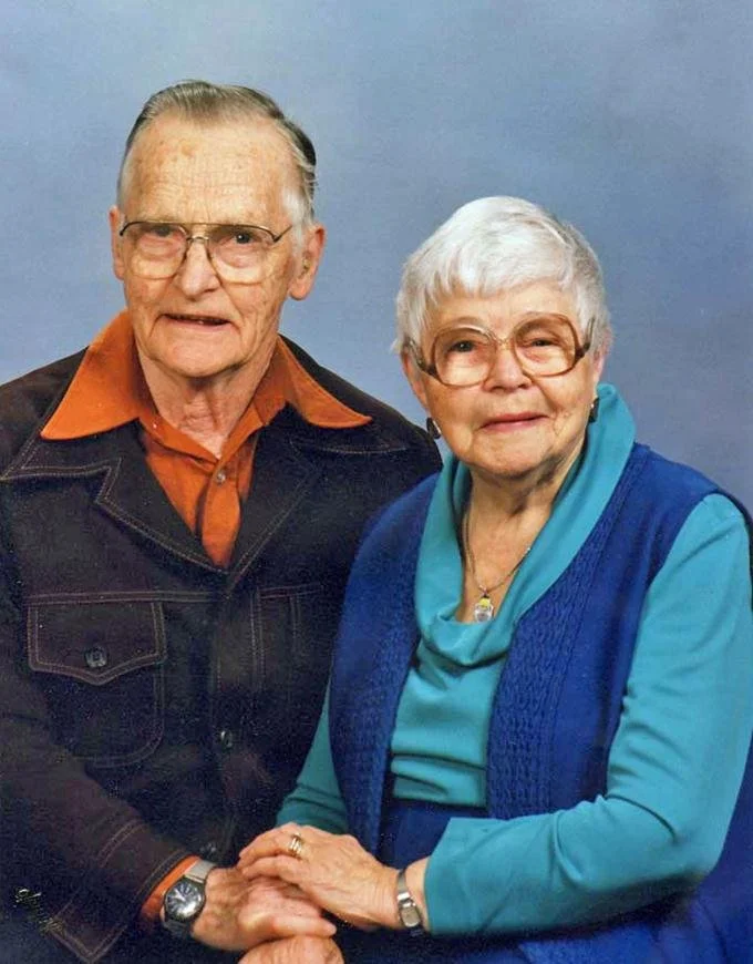 An elderly man and woman holding hands in front of a light blue background, both wearing glasses and smiling.