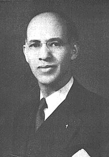 Black and white portrait of a bald man wearing glasses, a suit, and a tie.