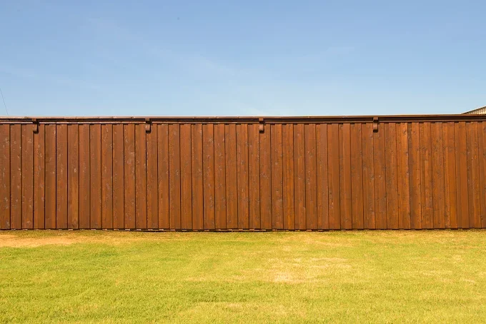 A tall wooden privacy fence with vertical planks and a flat top, set against a clear blue sky, with a green lawn in the foreground.