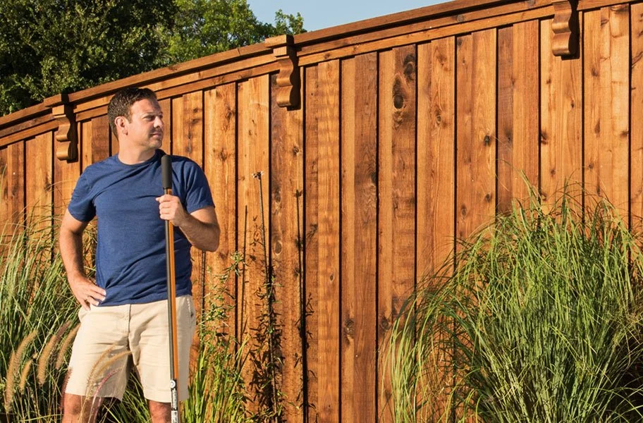 A man standing outdoors next to a wooden fence, holding a rake, wearing a blue t-shirt and beige shorts.