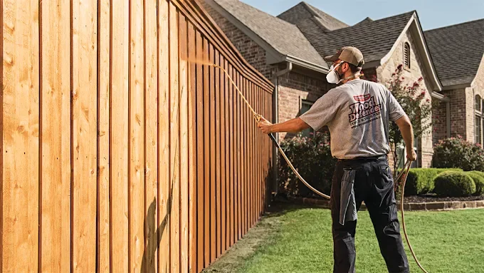 A man wearing a mask and cap is power washing a wooden fence in a backyard near a house.