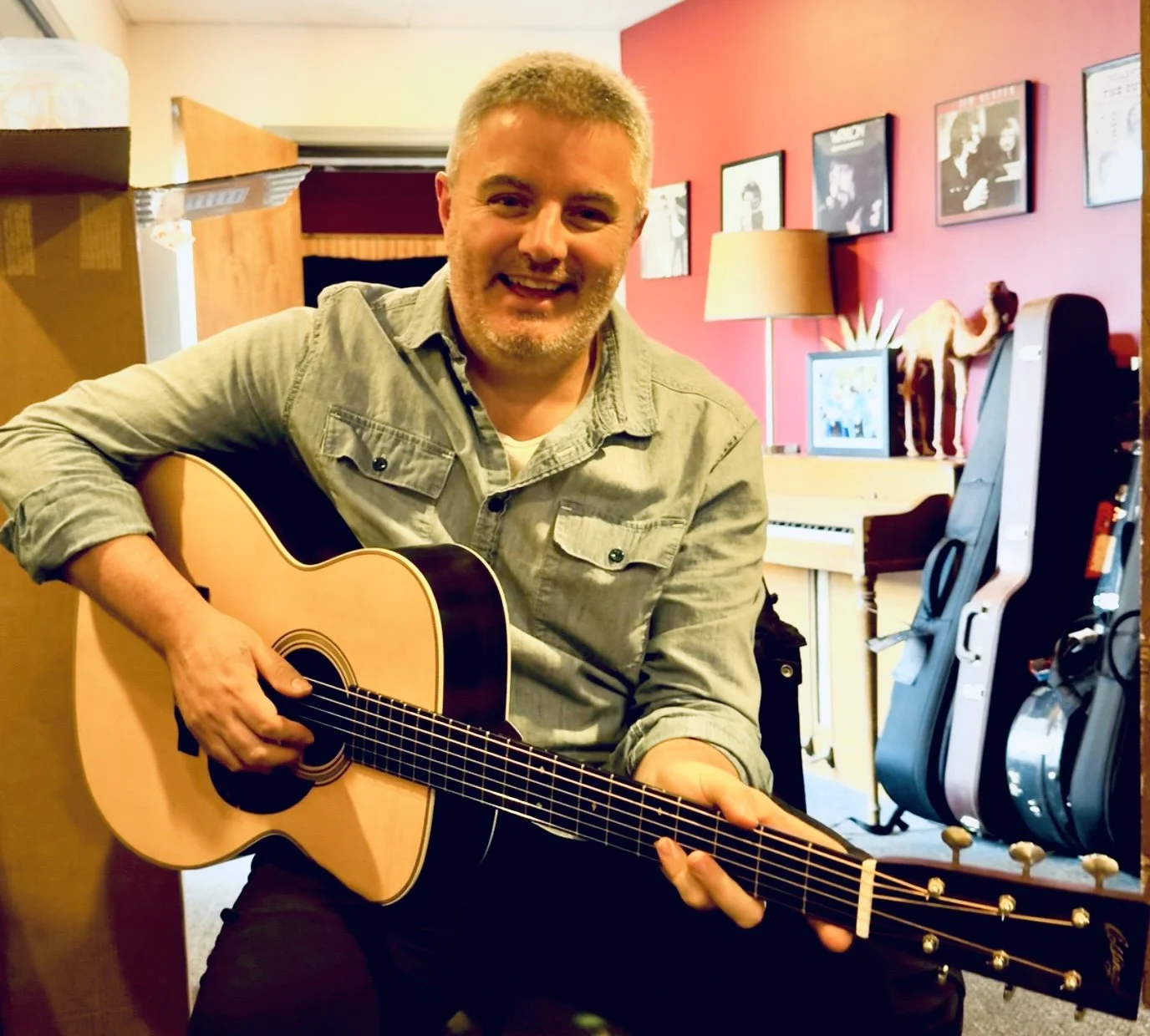 A man smiling and playing an acoustic guitar in a cozy room with red walls, framed photos, a lamp, a potted plant, and guitar cases in the background.