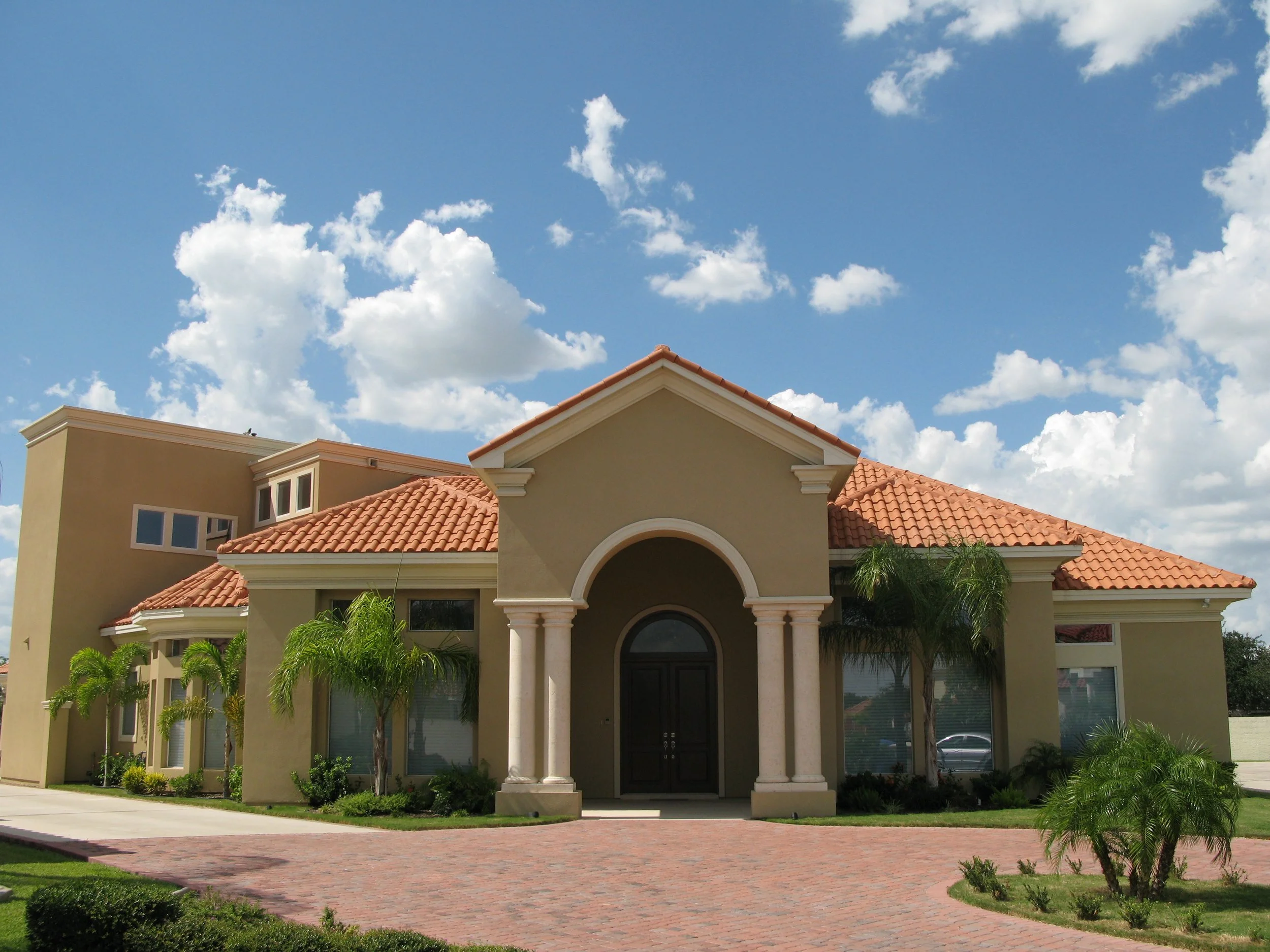 A large, elegant house with a tile roof, columns at the front entrance, and palm trees in the yard, under a partly cloudy sky.