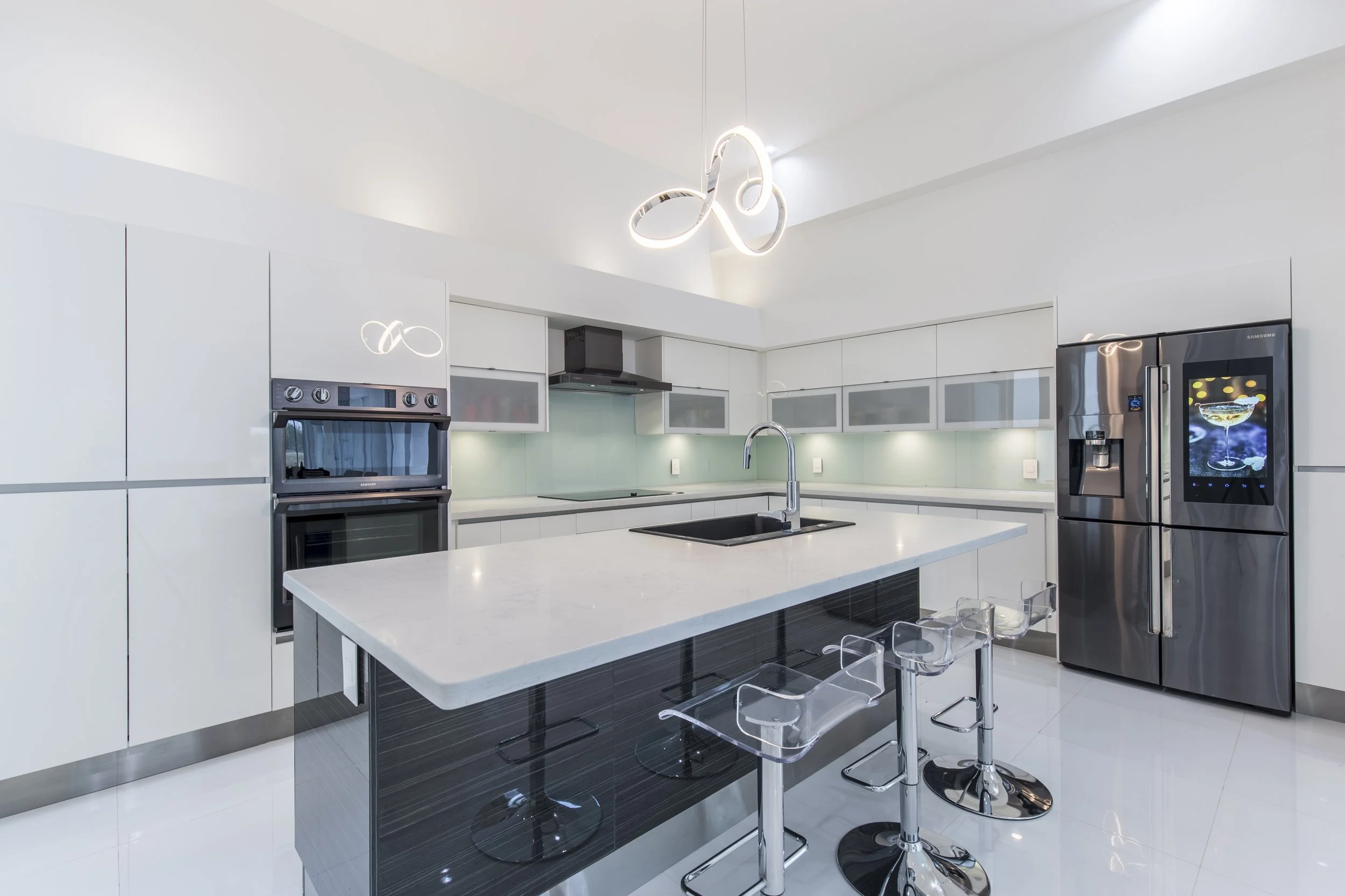 Modern white kitchen with a central island, stainless steel appliances, bar stools, and contemporary lighting fixtures.