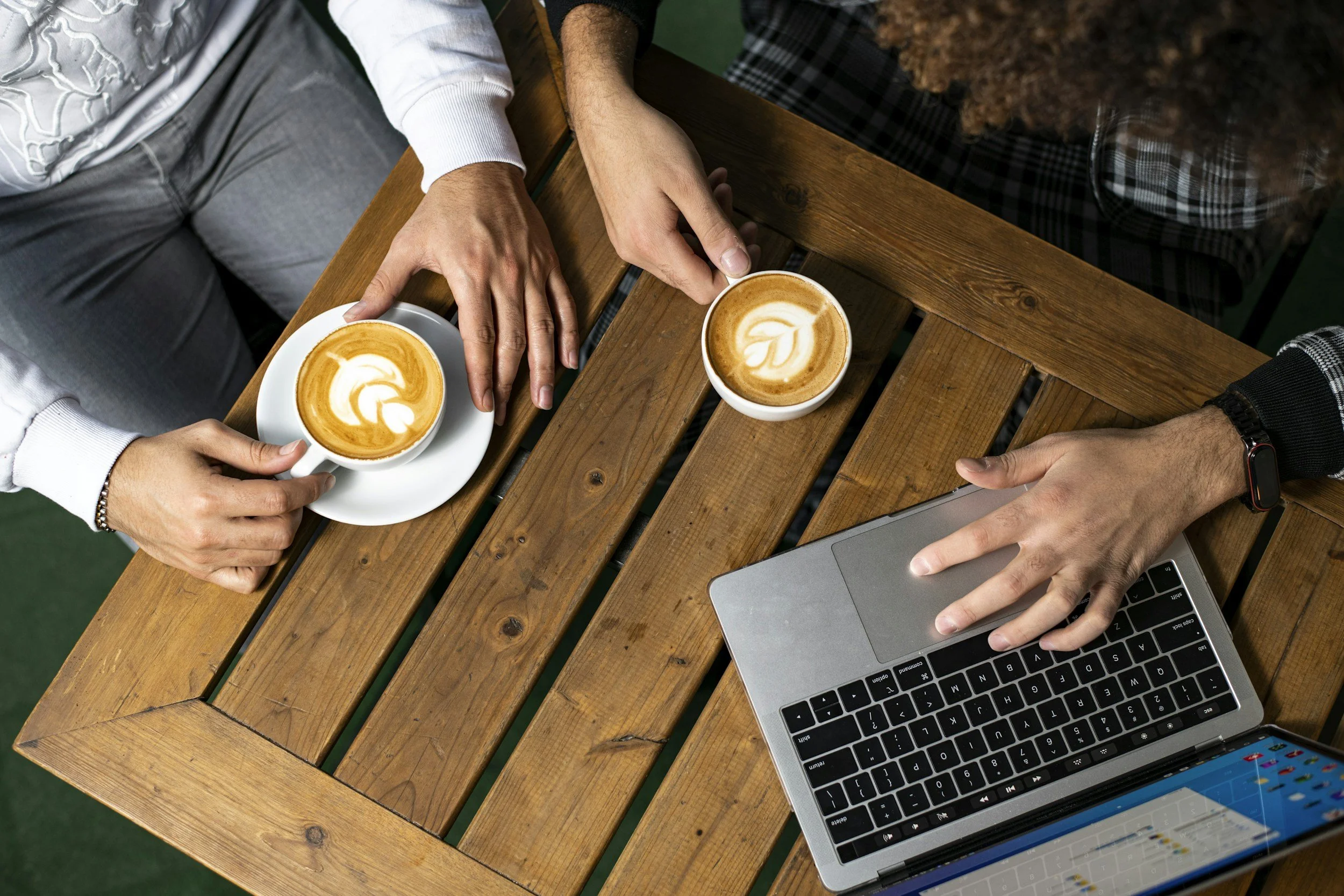 Two people sitting at a wooden table with two cups of coffee with latte art, one person using a silver laptop, and the other holding a coffee cup.
