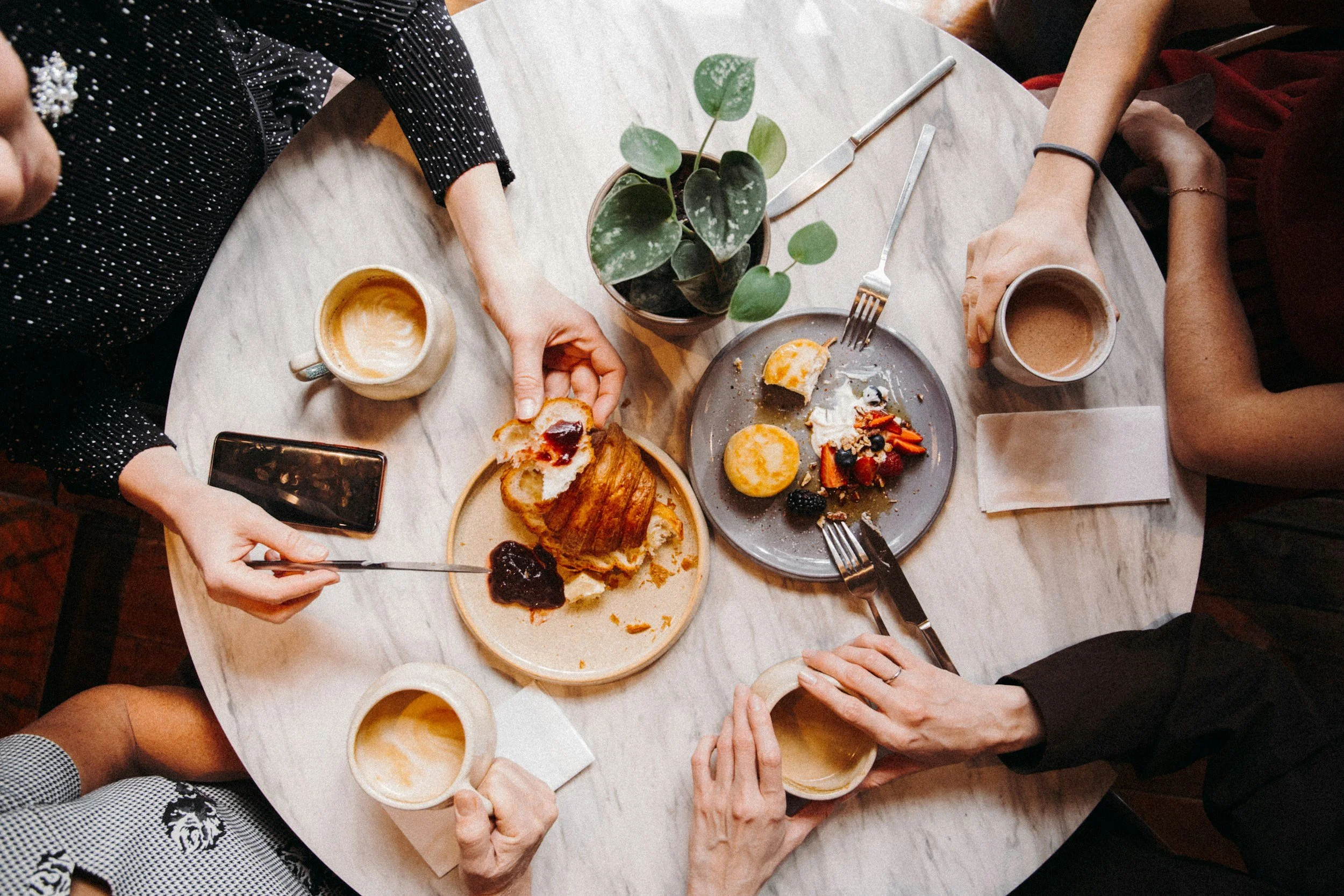 Four people sitting around a round table with desserts, cups of coffee or tea, and a potted plant. One person is reaching for a croissant with jam, others are holding cups.