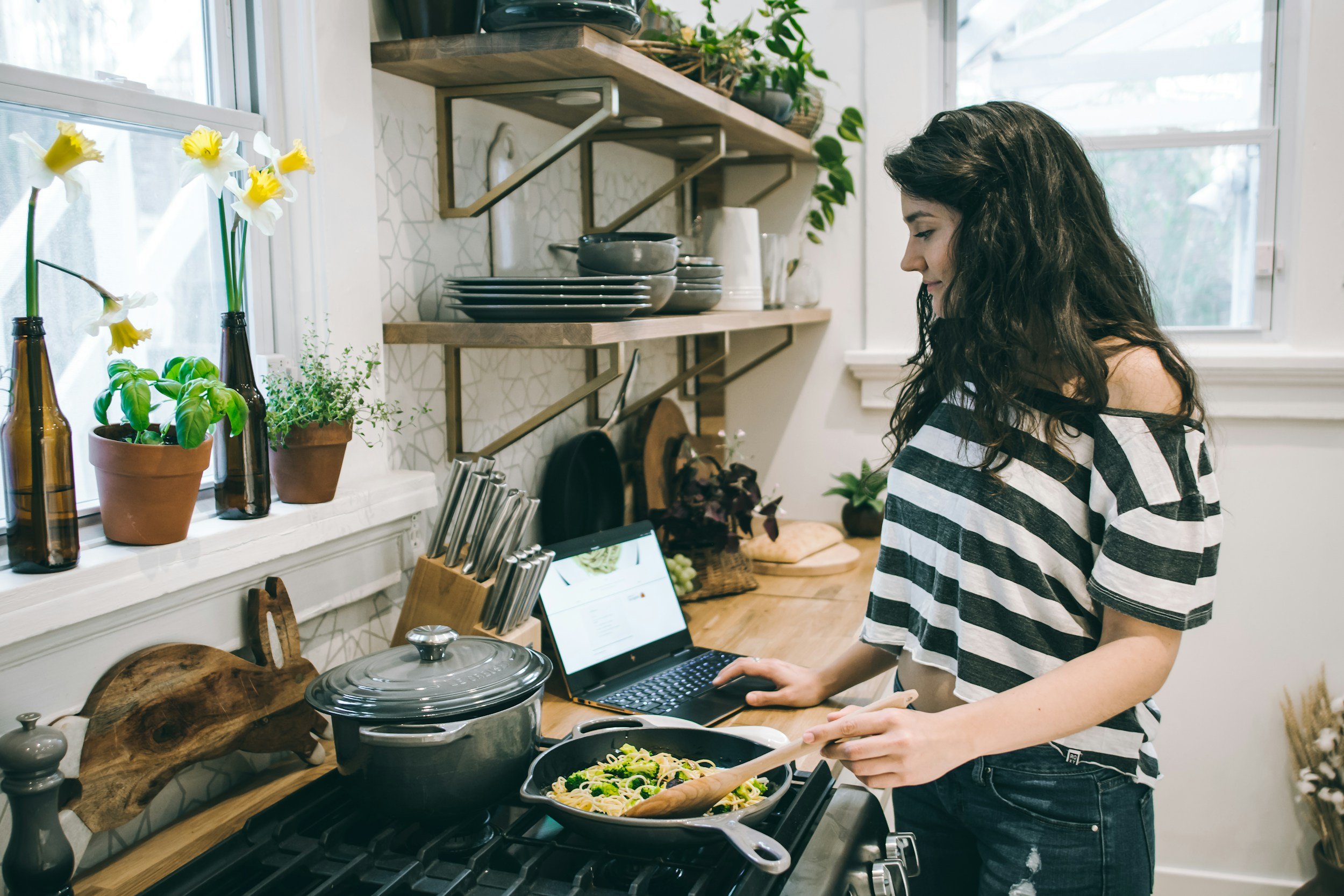 Young woman cooking pasta on stove while looking at a laptop in a bright, cozy kitchen with plants and open shelves.