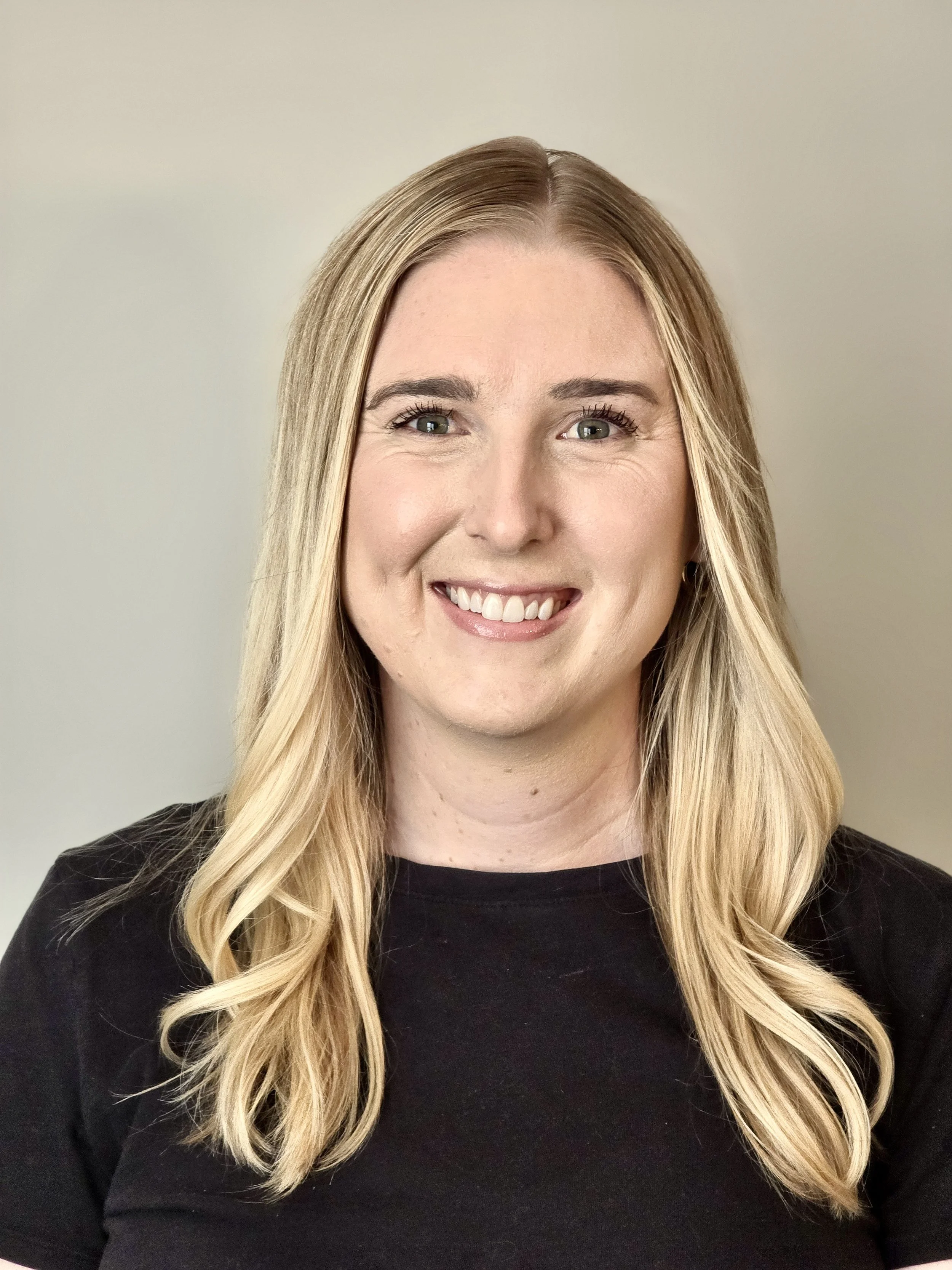 A woman with long blonde hair and a black shirt smiling, against a plain light-colored background.