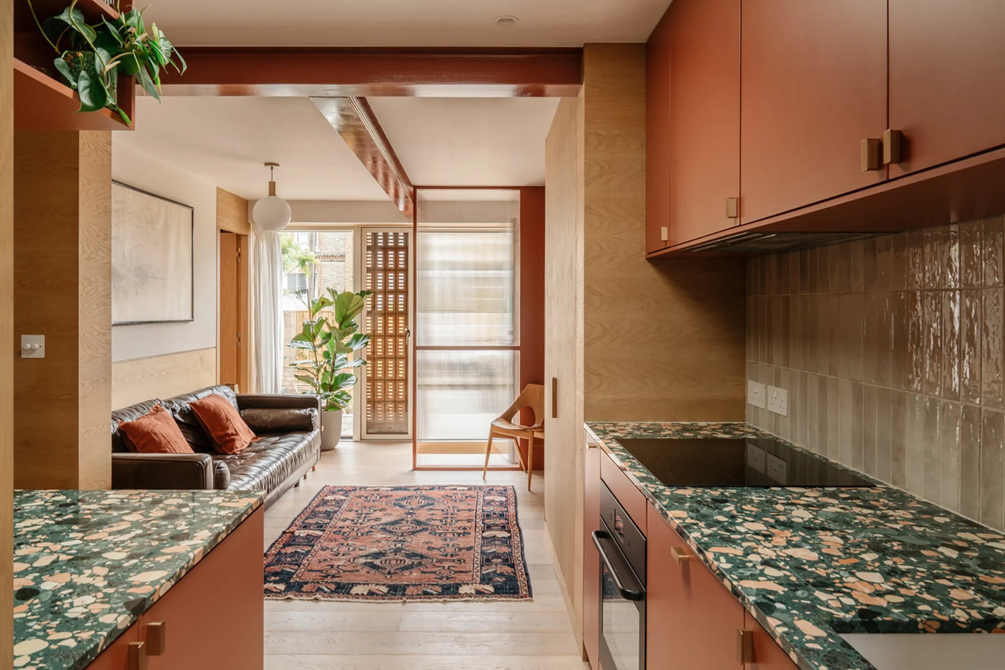 Open-concept living room and kitchen with wooden cabinets, green and cream granite countertops, and a leather sofa with orange pillows, large windows allowing natural light, potted plants, a patterned rug, and a sliding door with a wooden lattice.