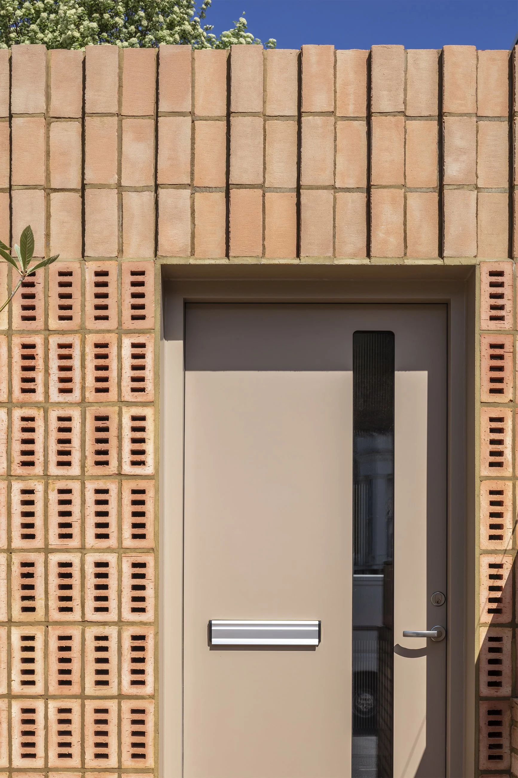 A beige door with a vertical black window and a metallic handle, set into a pink brick wall with evenly spaced brick patterns and a small plant leaf visible on the left, under a blue sky with some white clouds and green foliage in the background.