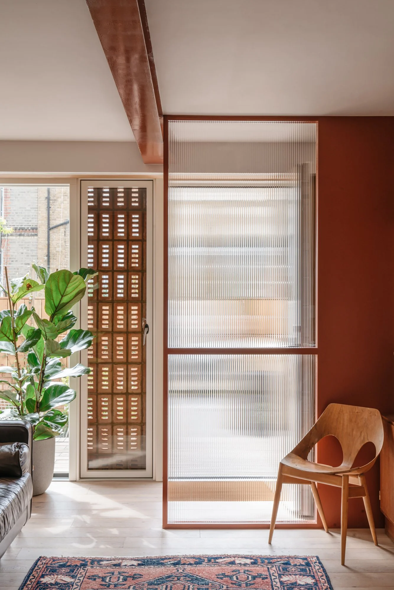 Interior of a modern living space with a large green indoor plant, a wooden chair, a textured glass room divider, and a door with decorative vented panels, sunlit through large windows.