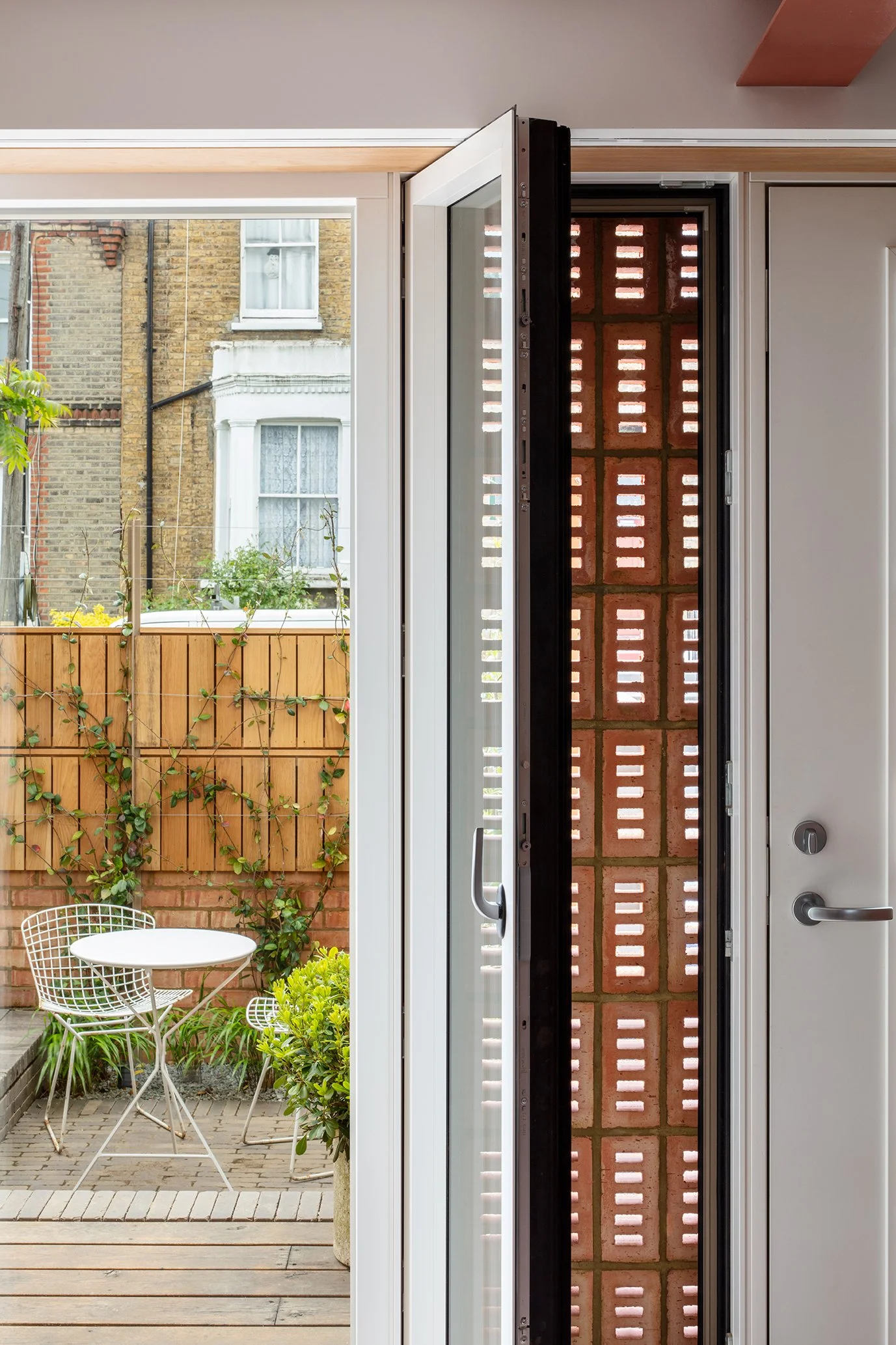 View from interior of a door opening to a small backyard patio with wooden flooring, potted plants, a metal table, and chairs, surrounded by a wooden fence with climbing plants, brick buildings, and windows in the background.