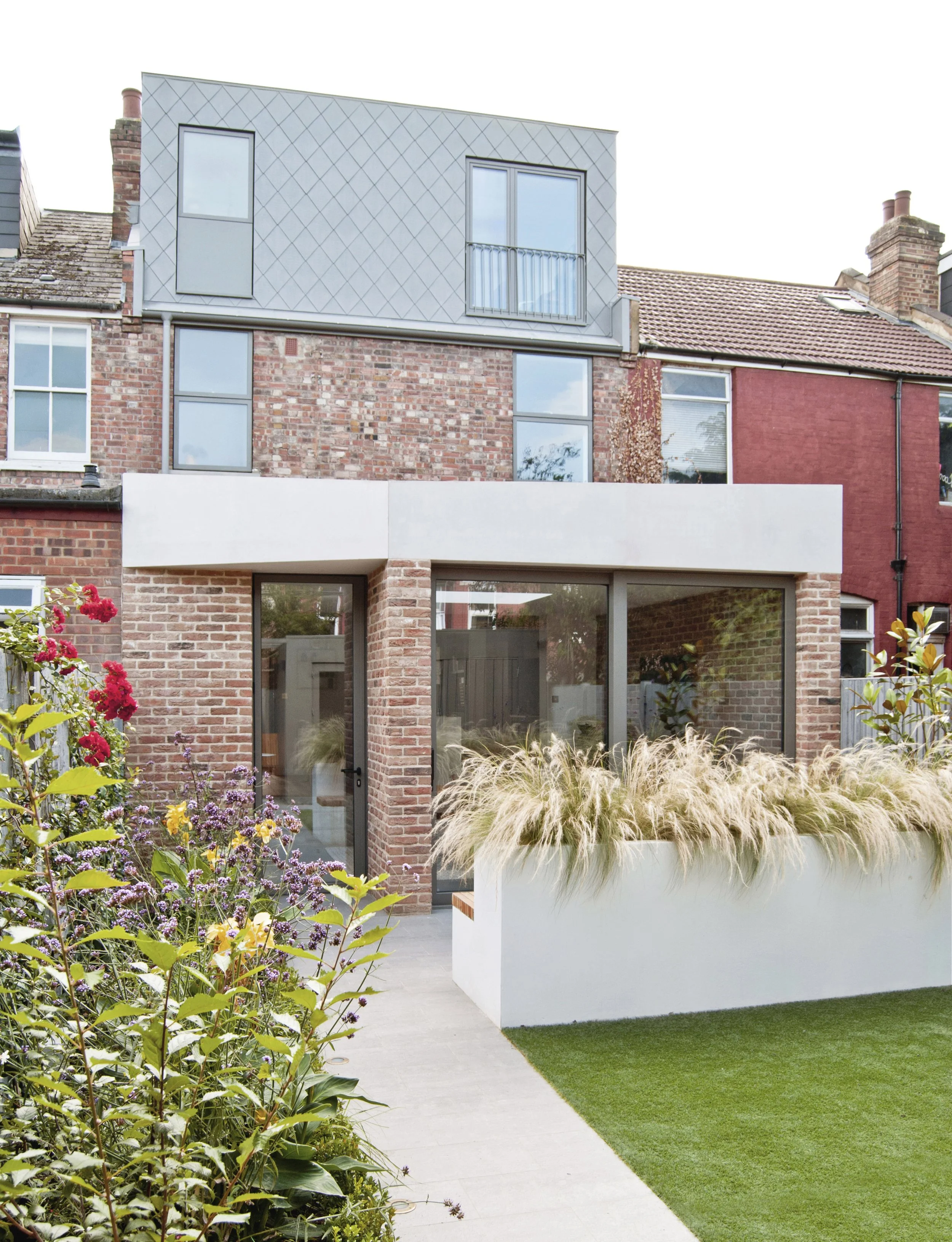 Modern residential building with brick and metal exterior, large glass windows, and a garden with flowering plants and ornamental grasses.