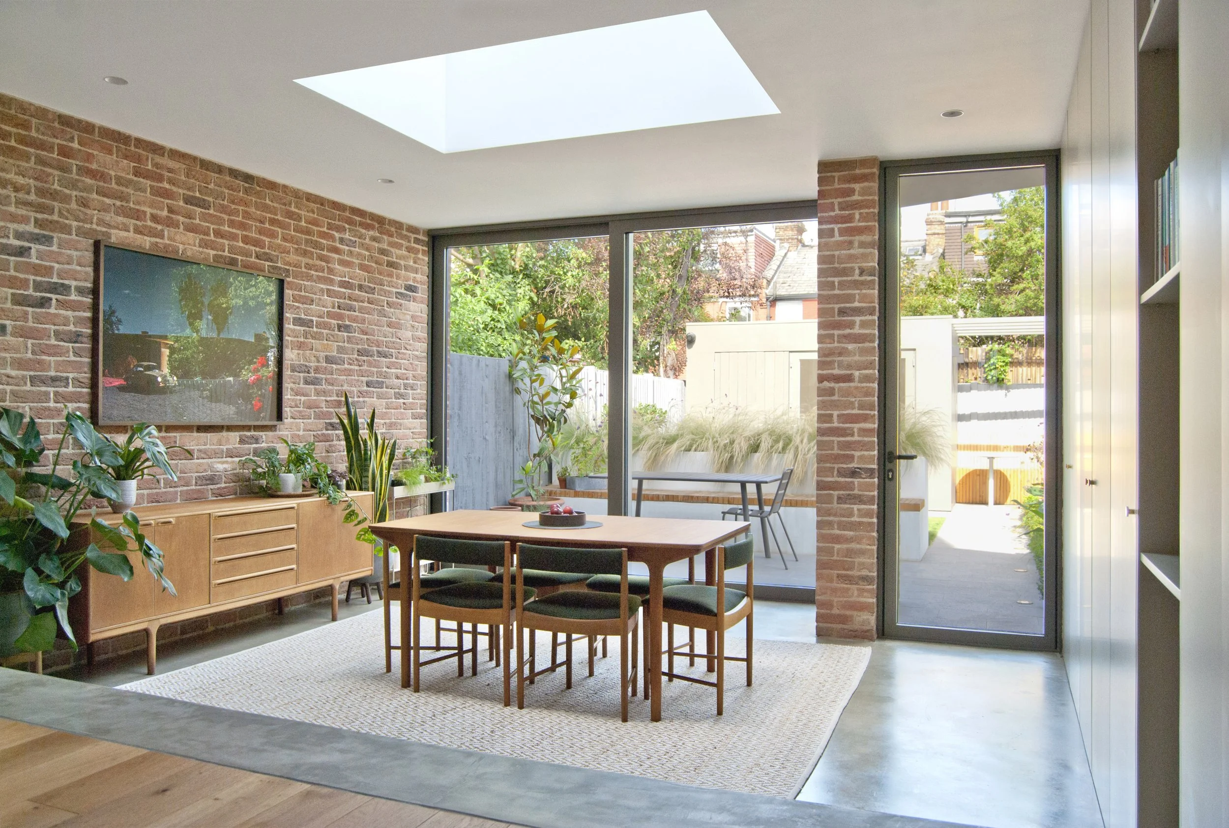 Modern dining room with brick walls, wooden dining table and six chairs, large sliding glass doors leading to backyard with patio, small outdoor table, green plants, and trees.
