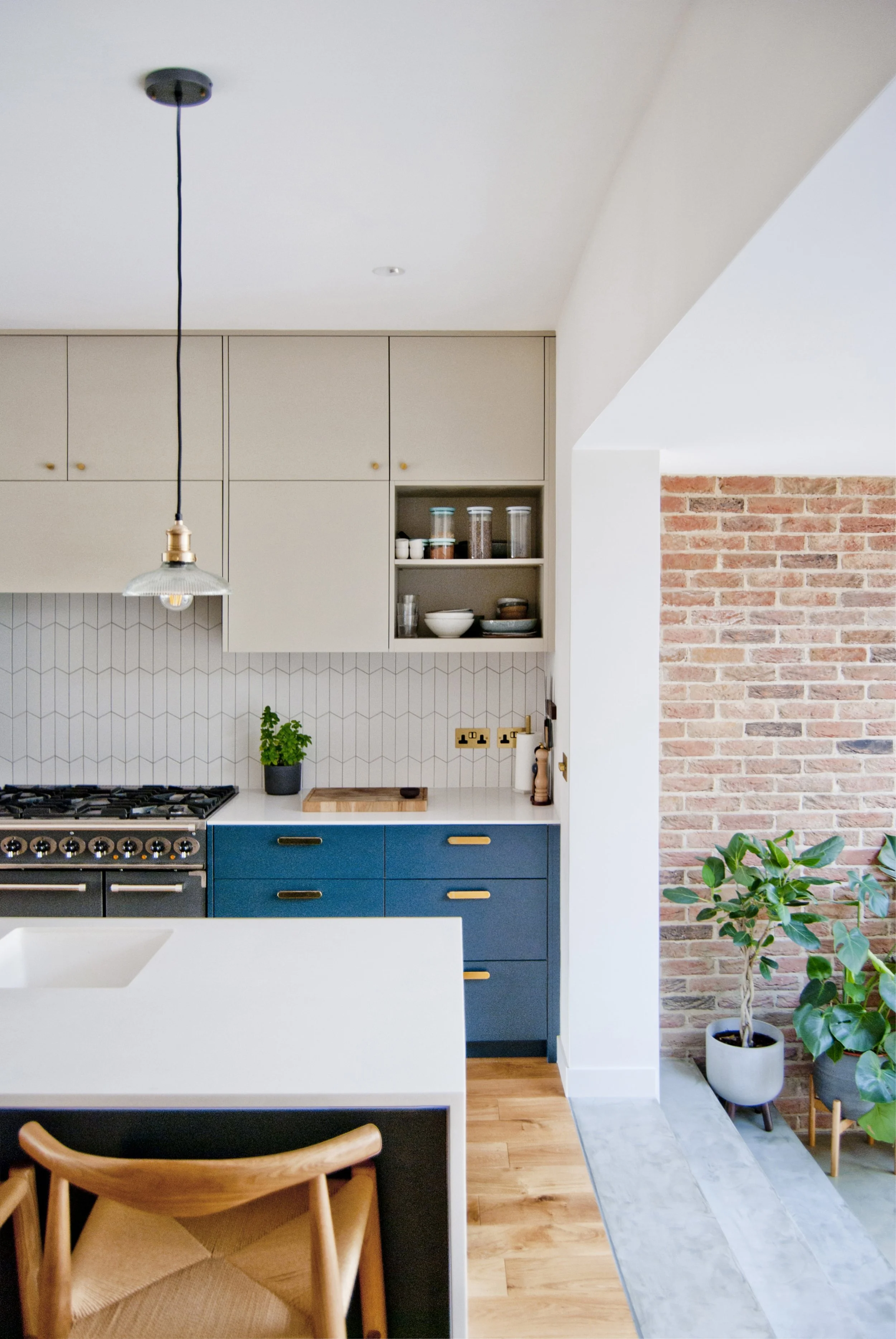 Modern kitchen with beige cabinets, blue lower drawers, a white countertop, a potted plant, open shelving with bowls and jars, and a brick accent wall.