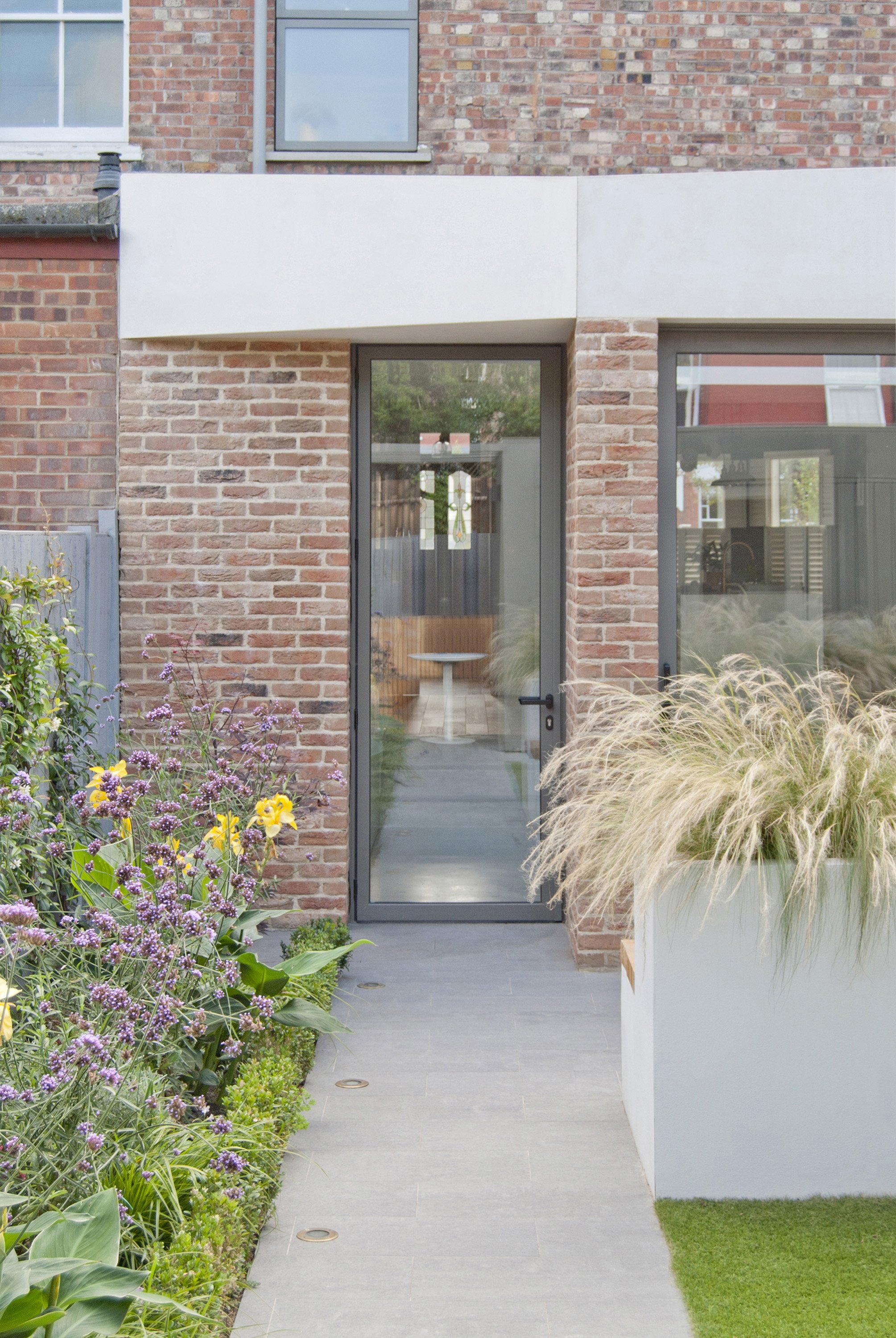 Modern house entrance with a glass door, brick walls, and outdoor plants including purple flowers and ornamental grasses, leading to an interior space visible through the glass door.