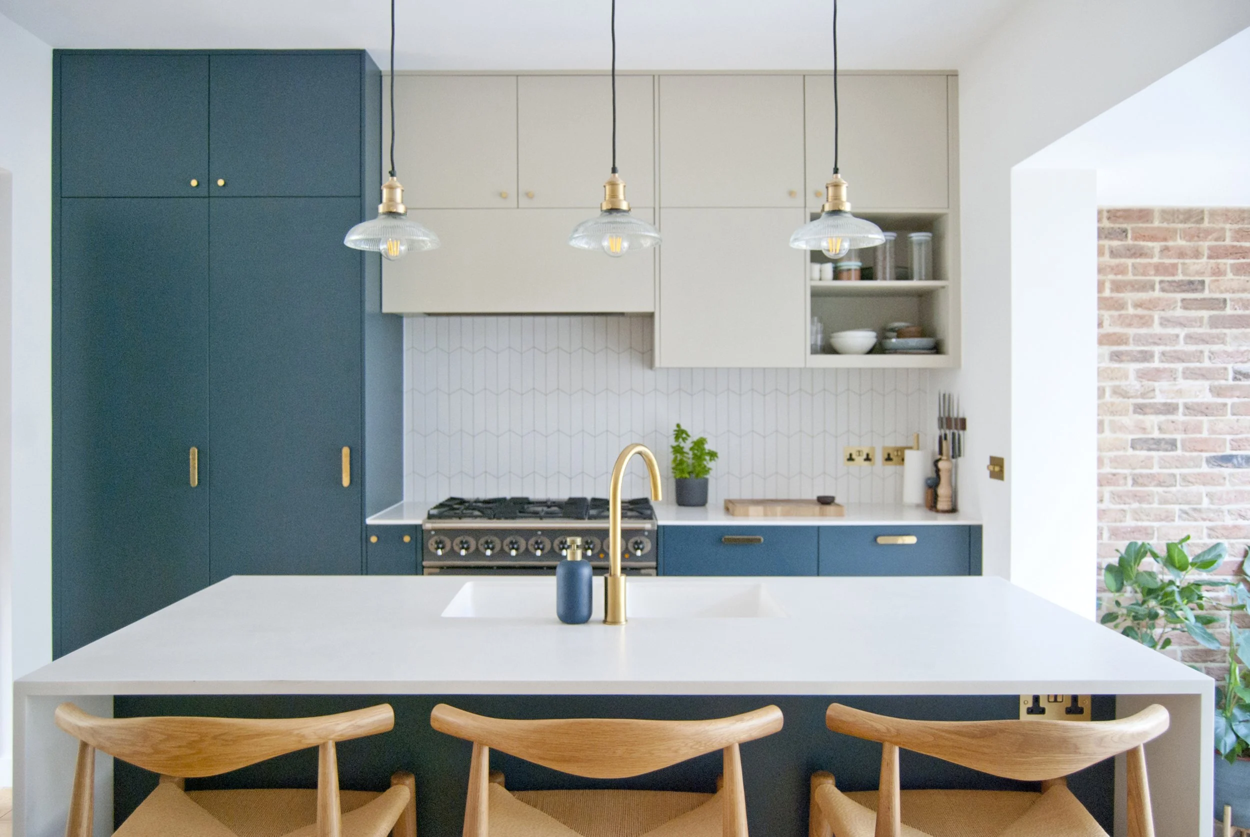 Modern kitchen with blue cabinets, beige upper cabinets, a white countertop island with a gold faucet, pendant lights, and a brick wall outside.