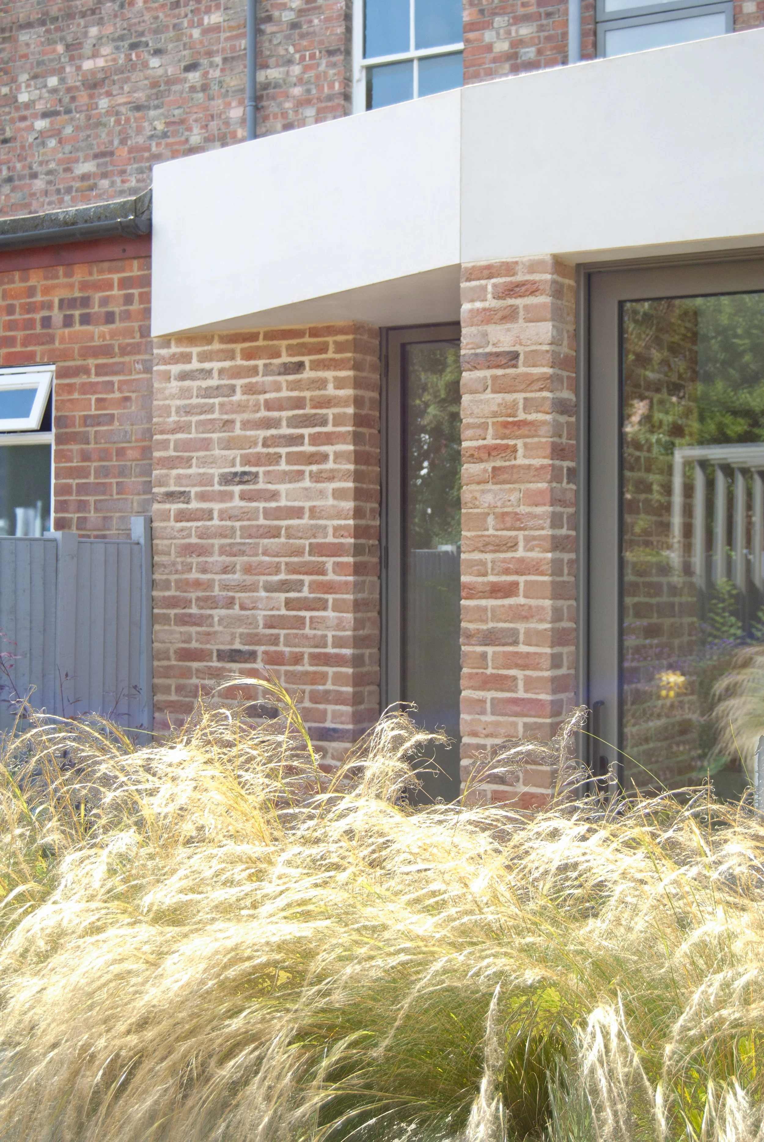 Exterior view of a modern brick house with large glass doors, a white overhang, and golden ornamental grasses in the garden.
