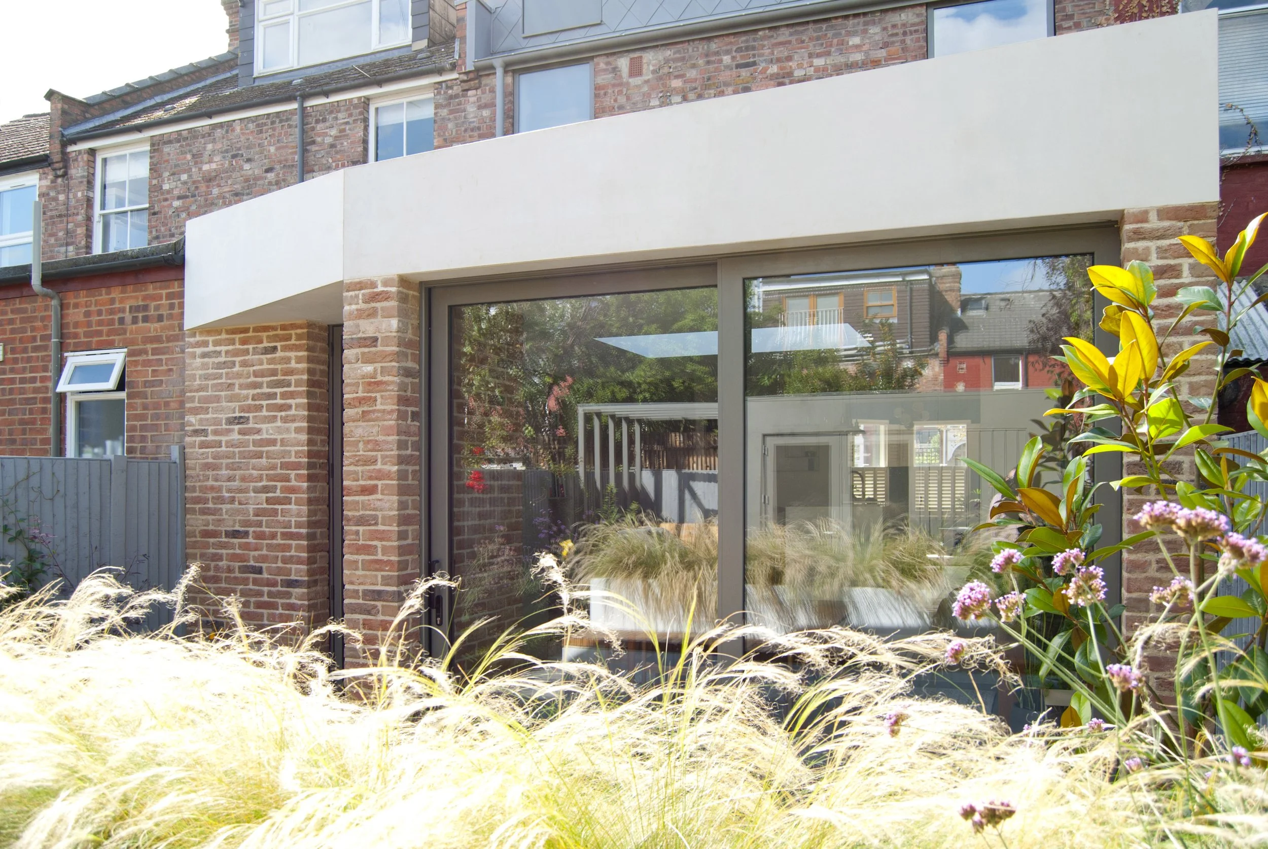 Modern brick house with large glass sliding doors, small garden in foreground with ornamental grass and flowering plants, neighboring brick houses visible in reflection.