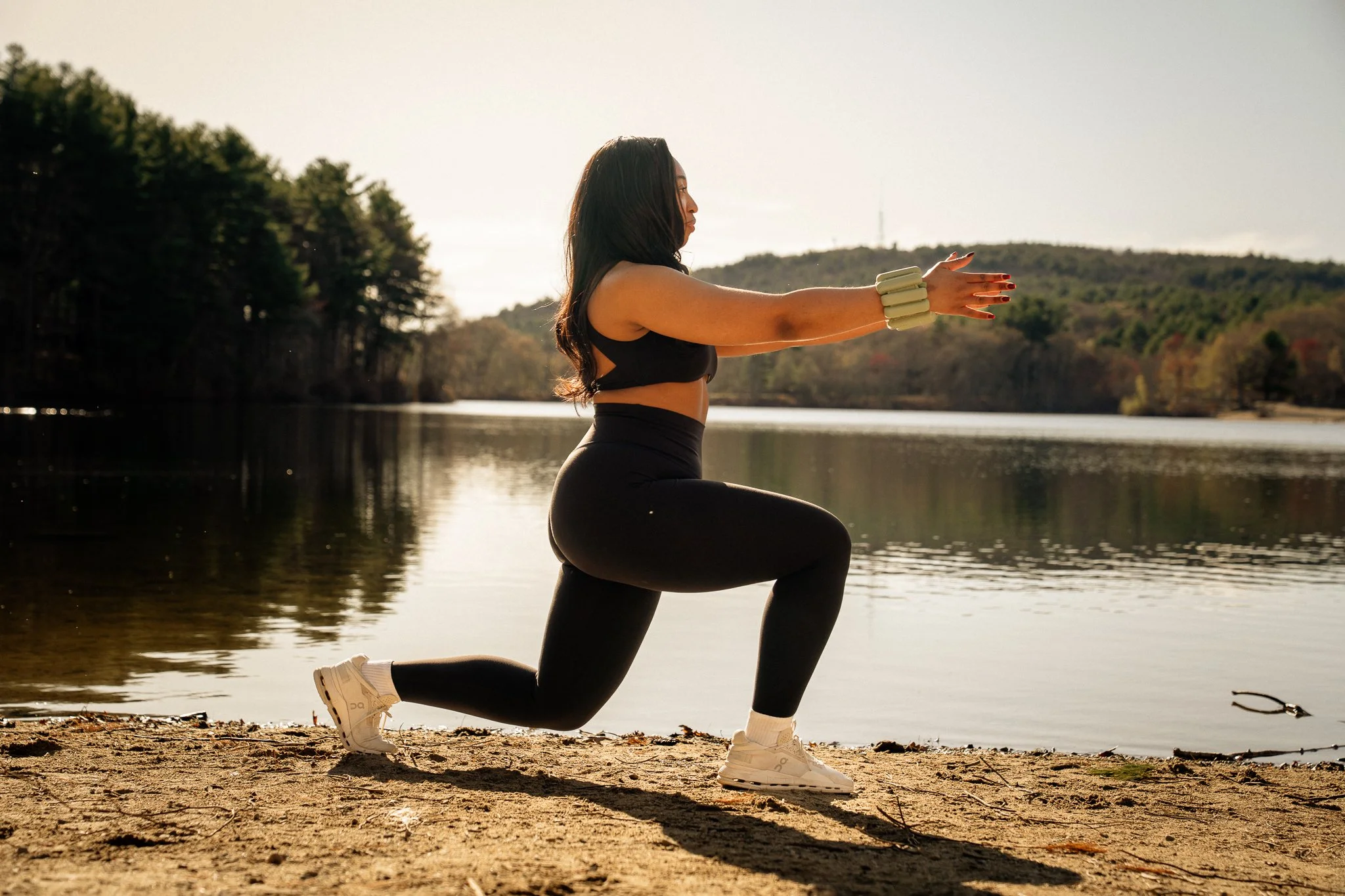 Woman practicing yoga on a lakeshore during sunset, kneeling in a lunge position with arms extended forward, wearing black athletic clothing and white sneakers.