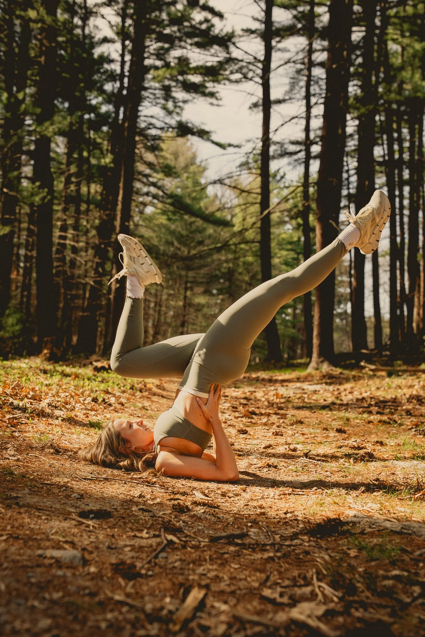 A woman practicing yoga outdoors in a forest, doing a shoulder stand pose with her legs extended upwards. She is lying on the ground on her back with her hands supporting her lower back, surrounded by trees and fallen leaves.