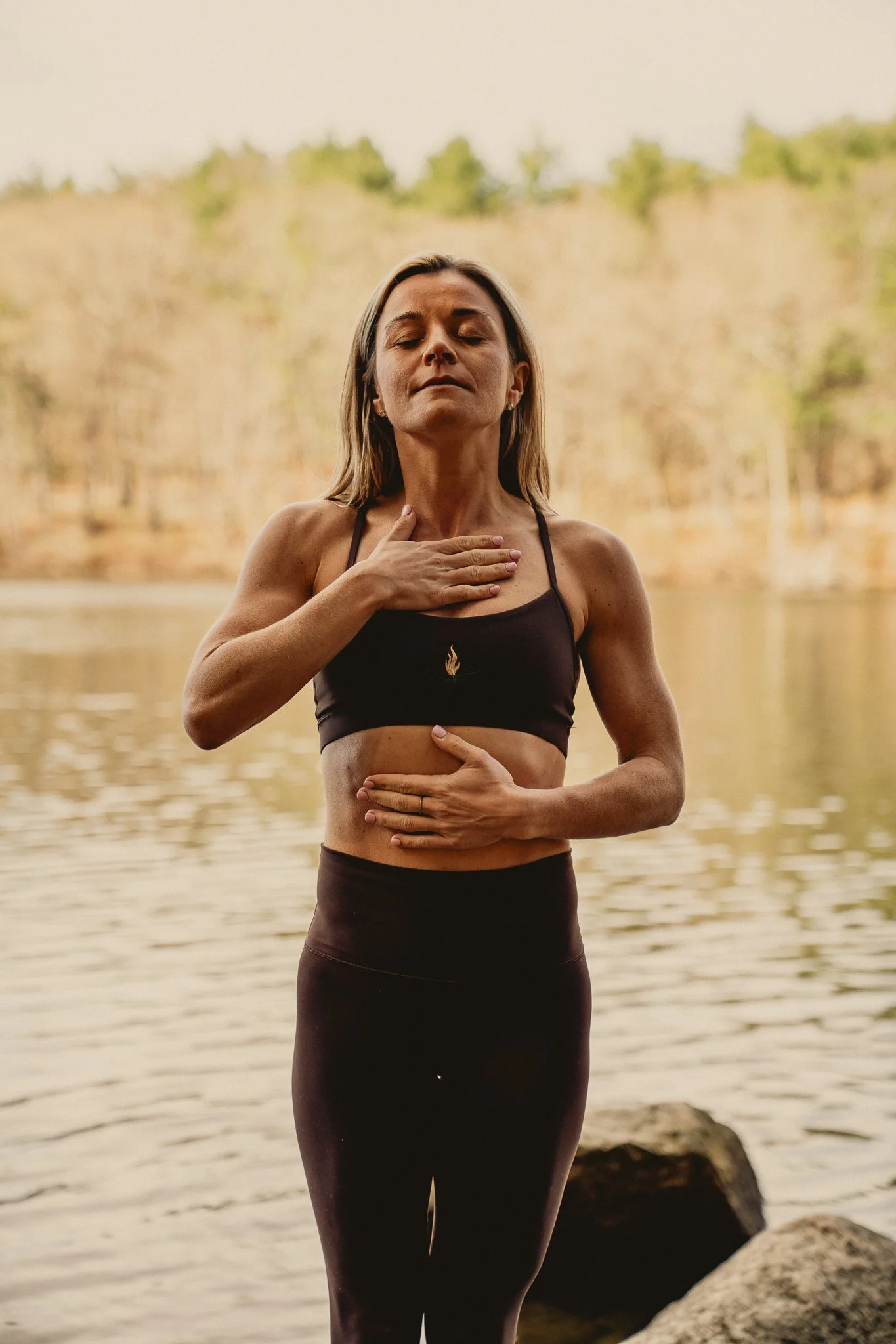 A woman practicing yoga or meditation by a body of water, with her eyes closed and one hand on her chest and the other on her stomach, during sunset or sunrise.