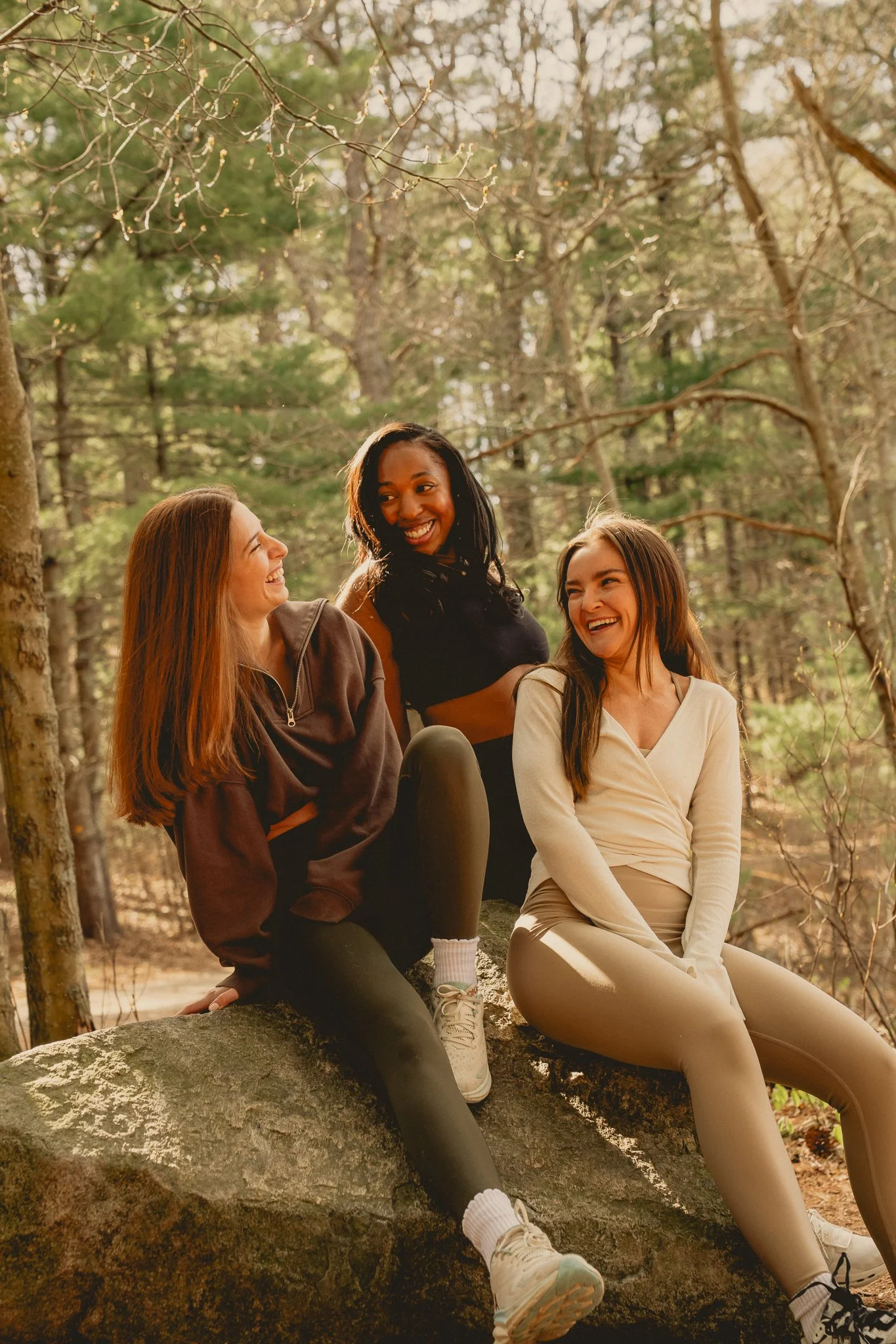 Three women sitting on a large rock in a forest, smiling and talking to each other, with trees and sunlight in the background.
