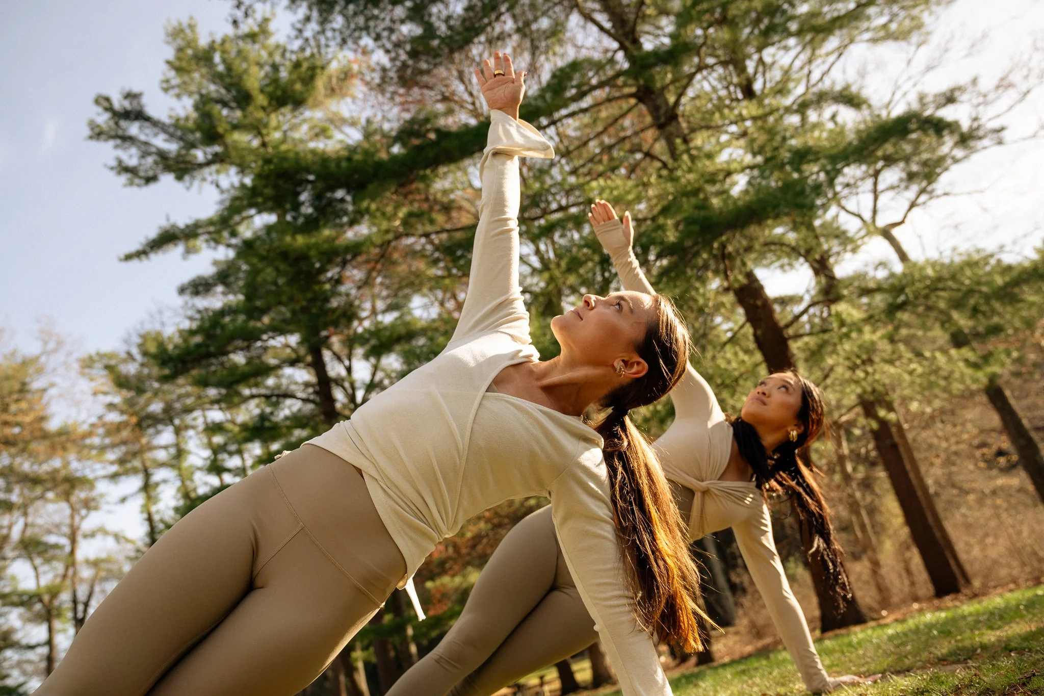 Two women practicing yoga outdoors in a park, performing a side stretch pose, with trees in the background.