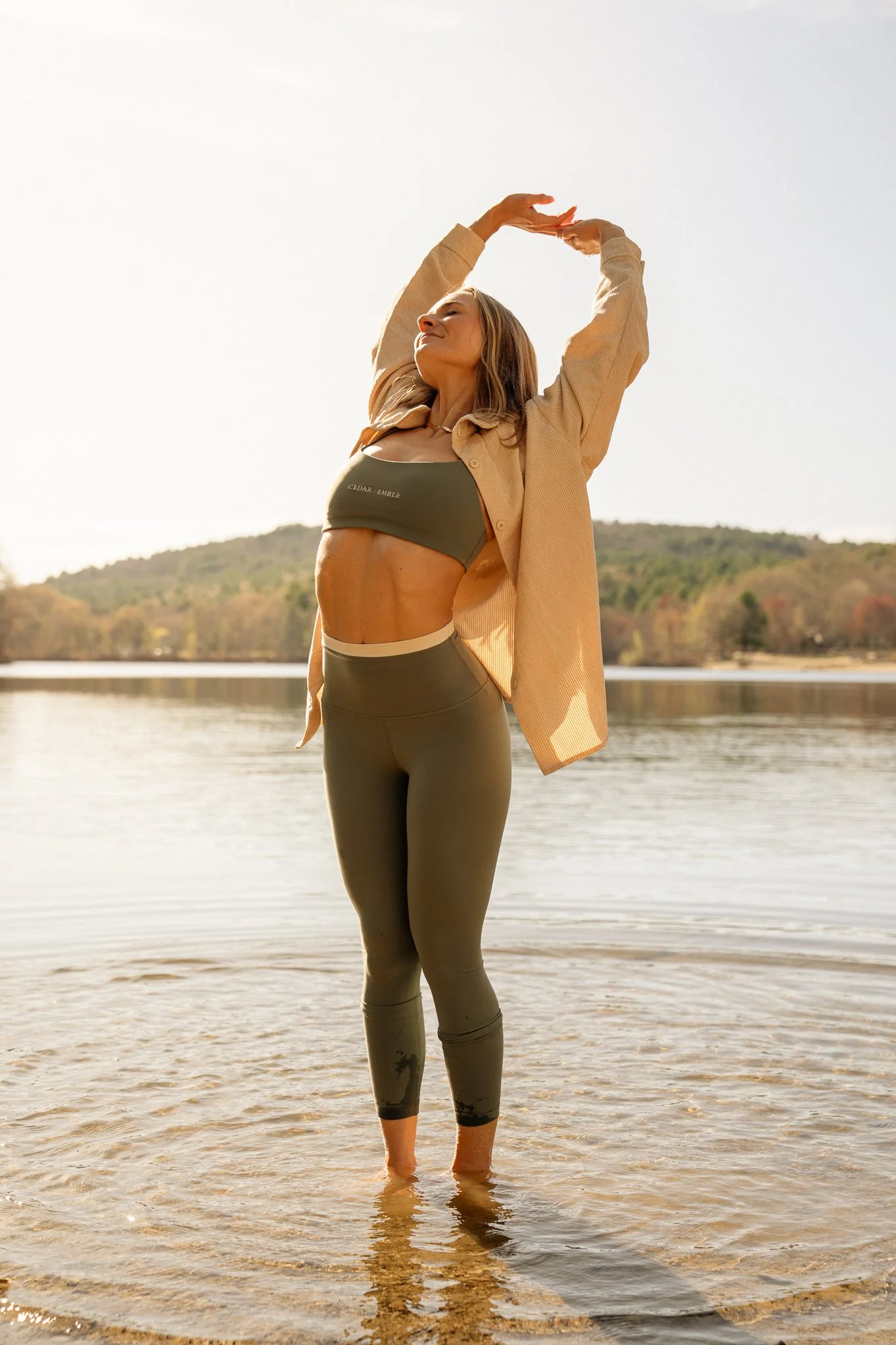 A woman standing in shallow water at a lake or river, stretching her arms above her head and smiling in the sunlight with trees and hills in the background.