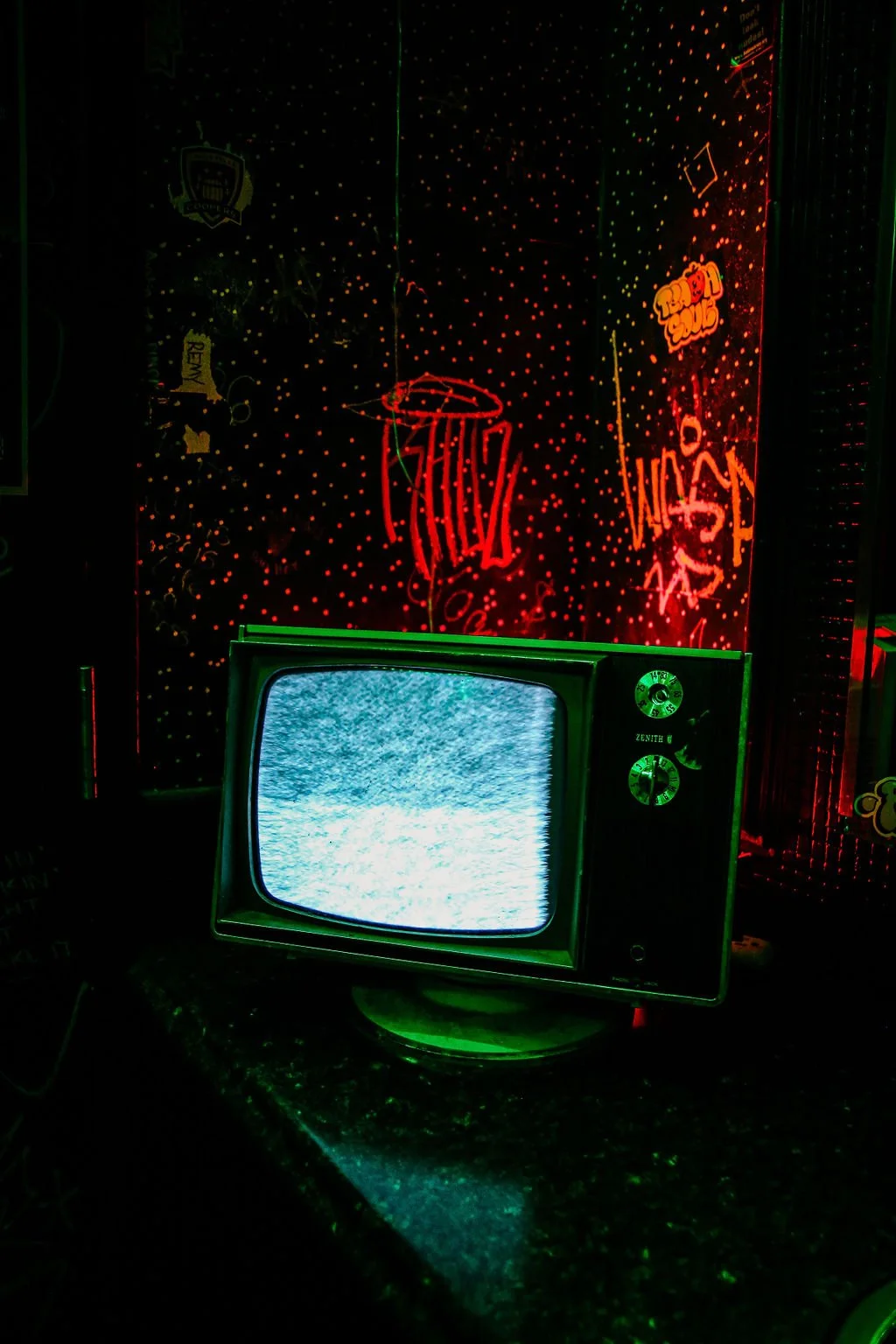 A vintage television displaying static, with neon signs and graffiti-style art on the wall behind it, illuminated in red and green lights.