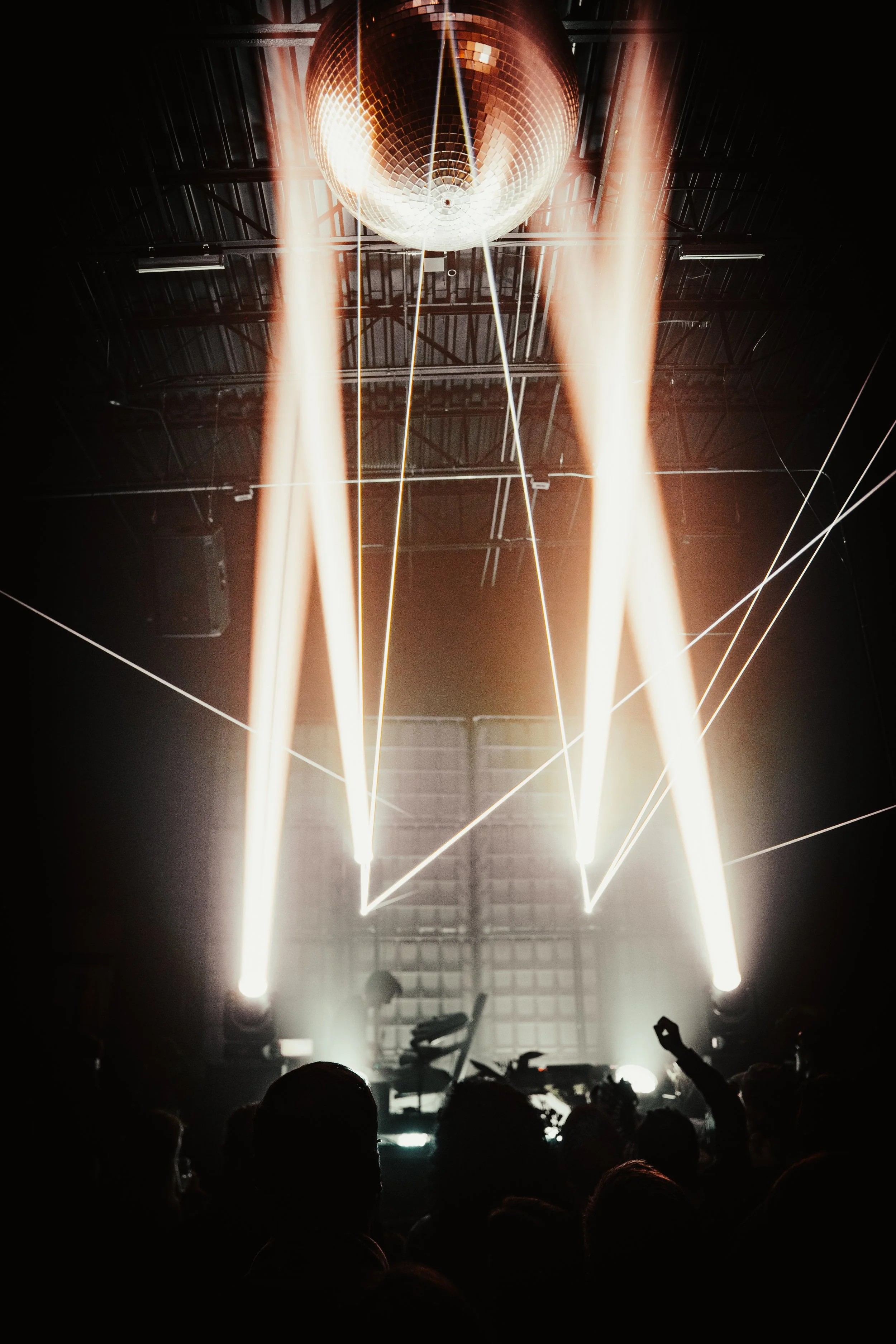 A dark concert scene with a large disco ball hanging from the ceiling, illuminated by four bright white lights casting beams downward. An audience is visible in silhouette at the bottom of the image, with some people raising their hands.