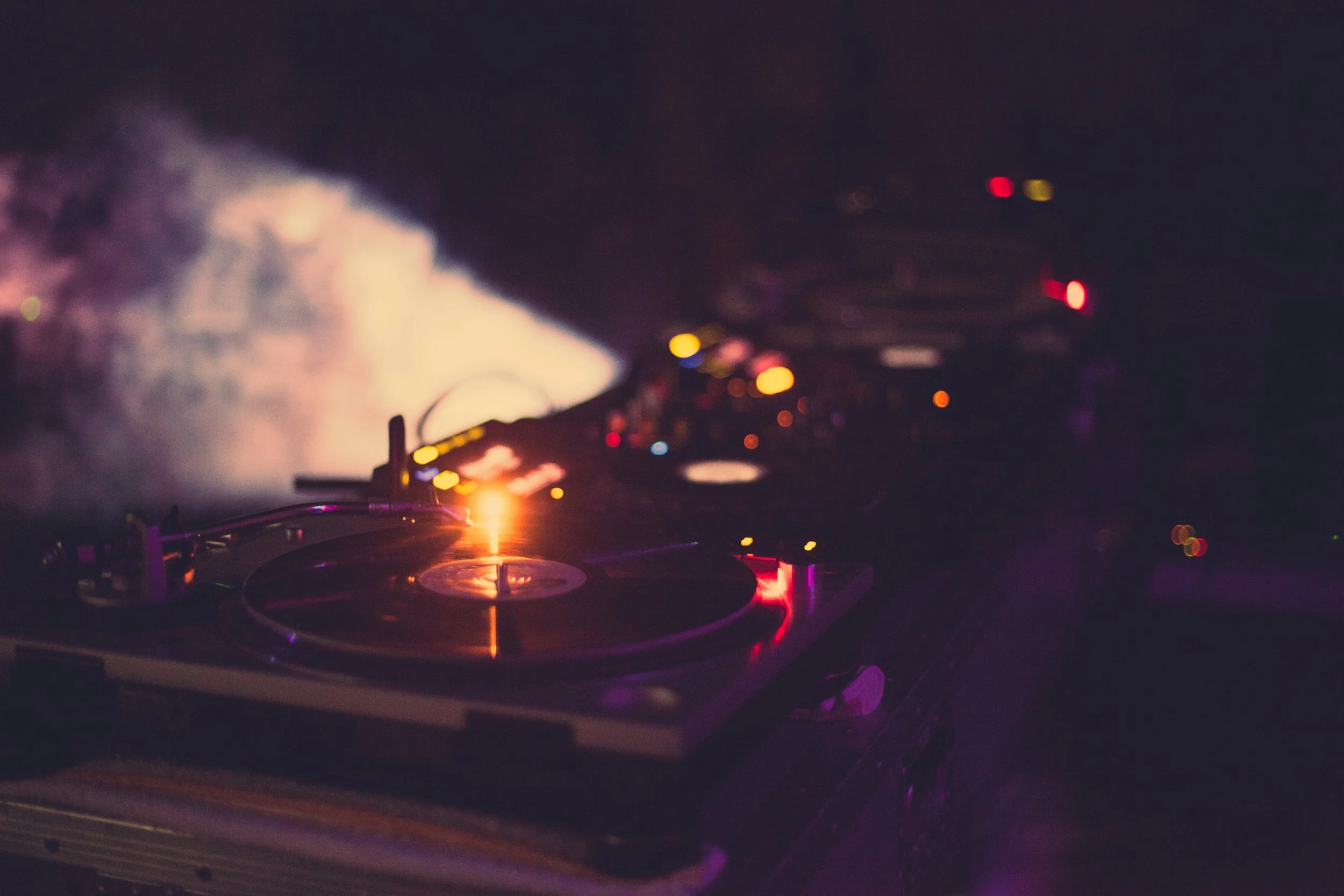 A DJ turntable setup with colorful blurry lights and smoke, creating a vibrant nightclub or party atmosphere.