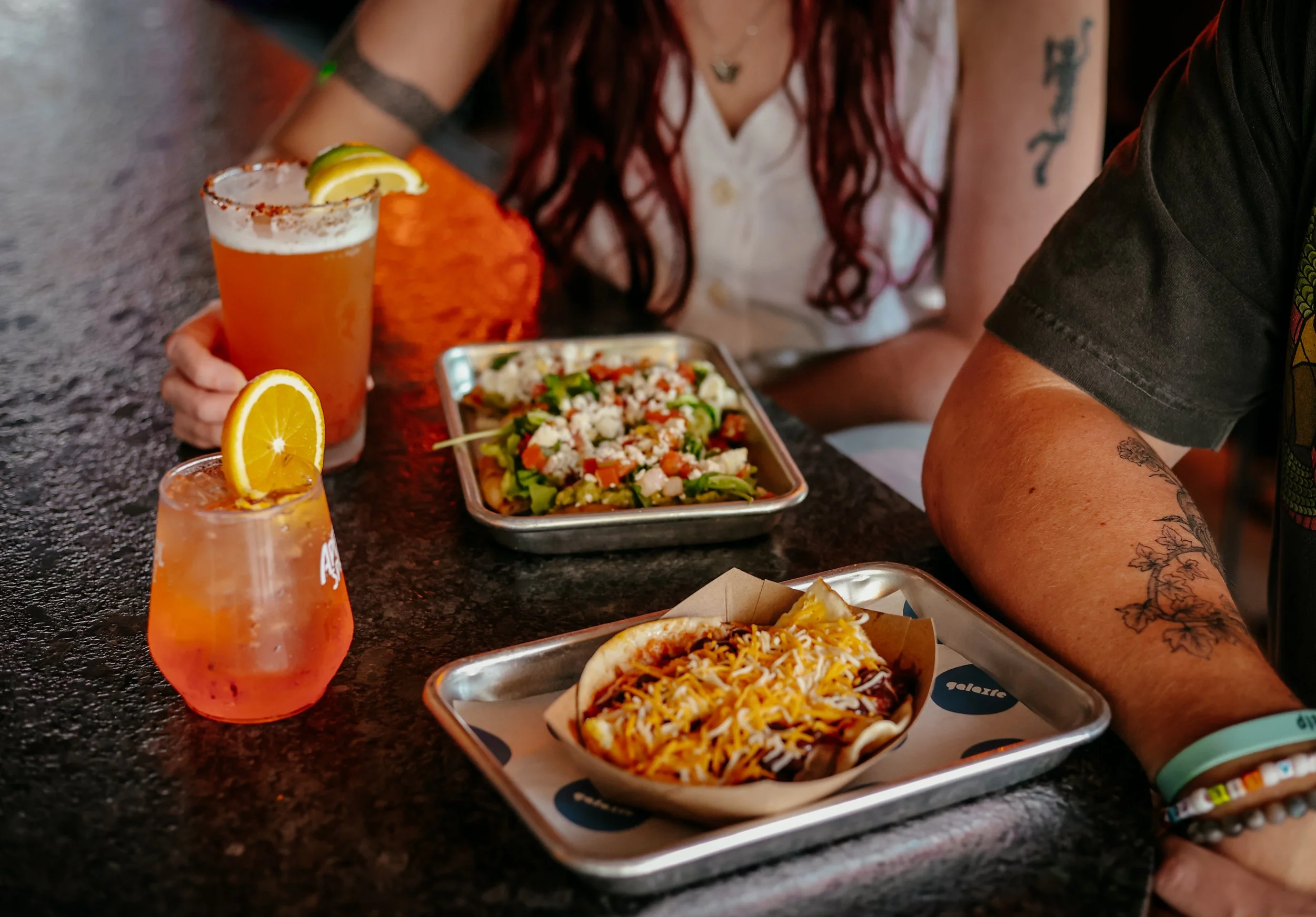 A table with a drink garnished with lemon and chili powder, two food dishes with cheese, and a woman with red hair, tattoos, and wearing a white shirt, sitting next to a man with a black shirt.