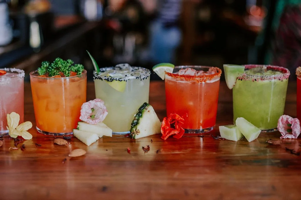 Line of five colorful cocktails garnished with fruits and herbs on a wooden table.