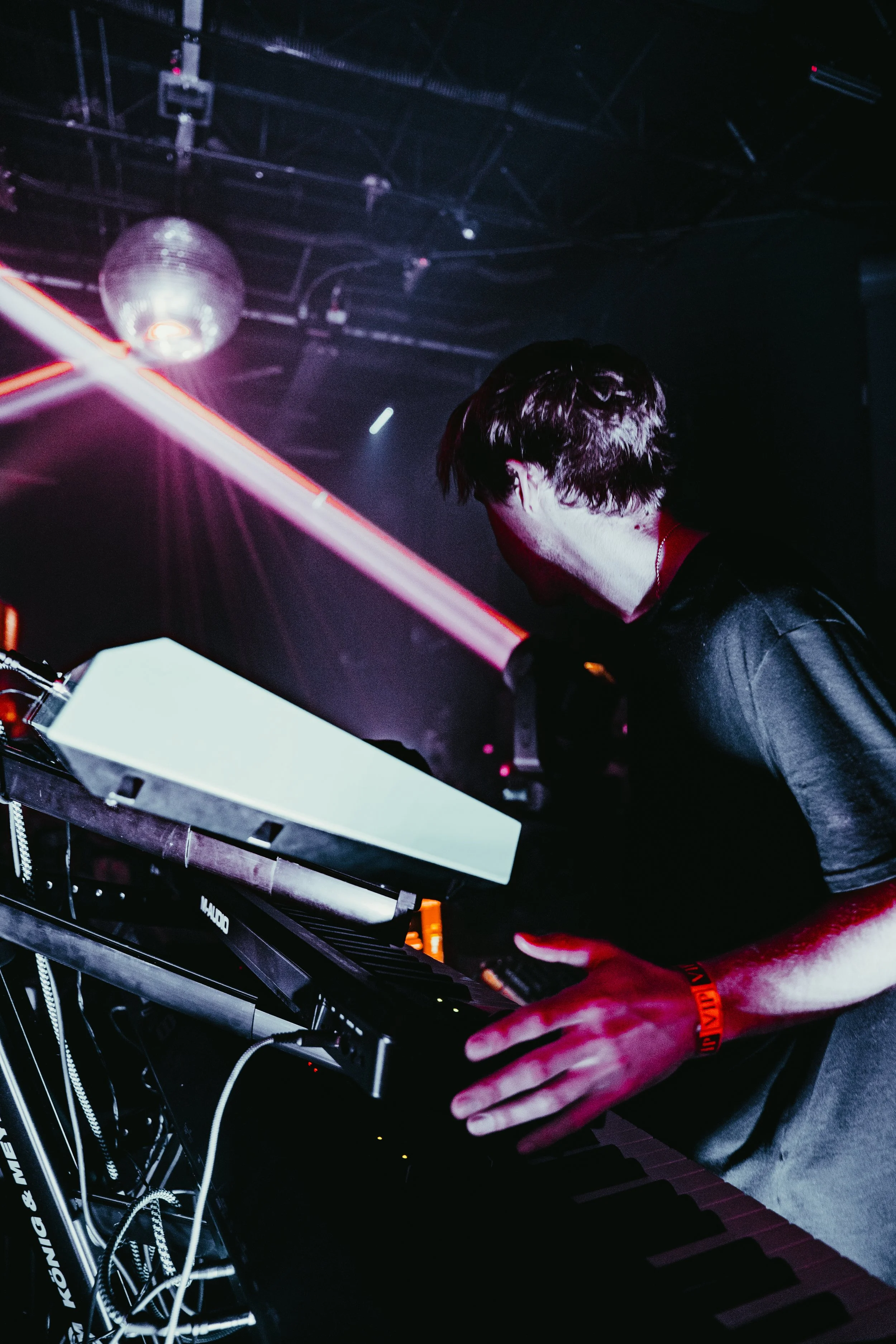 A person with dark, styled hair wearing a black shirt, playing a keyboard in a dark club or concert setting with a disco ball and various lighting.
