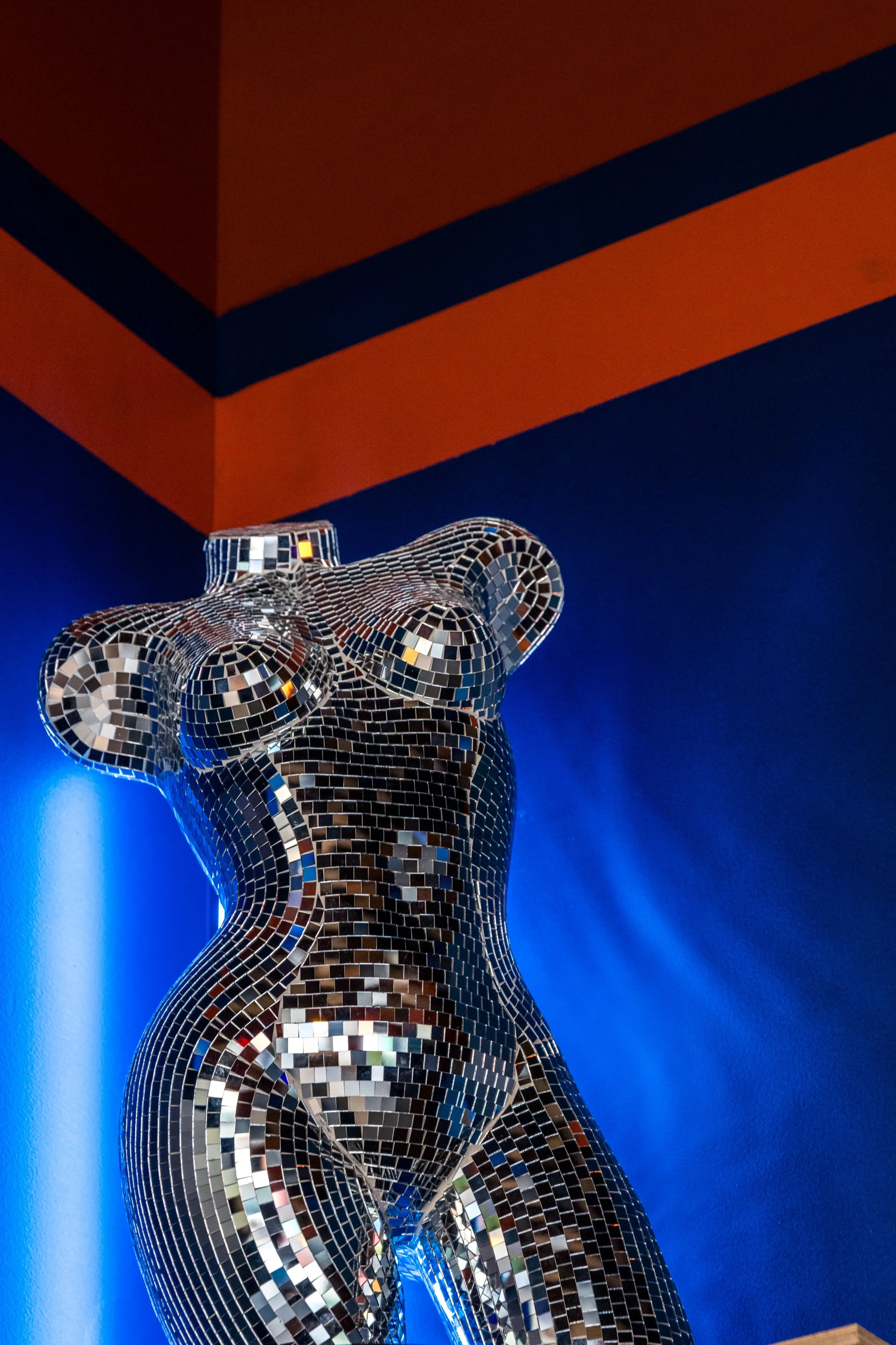 A reflective, mosaic-covered sculpture of a woman’s torso and arms, illuminated with blue lighting against a dark blue wall and orange ceiling with black stripes.