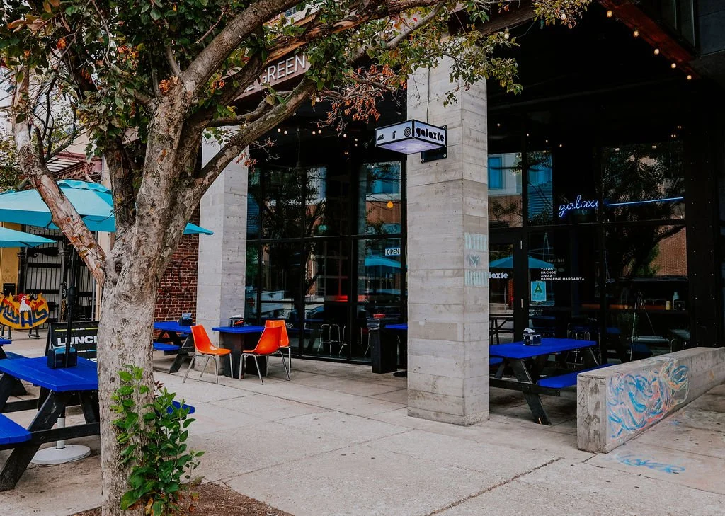 Exterior view of a modern indoor-outdoor restaurant or cafe with blue tables and orange chairs, a tree in the foreground, and large glass windows reflecting the street scene.