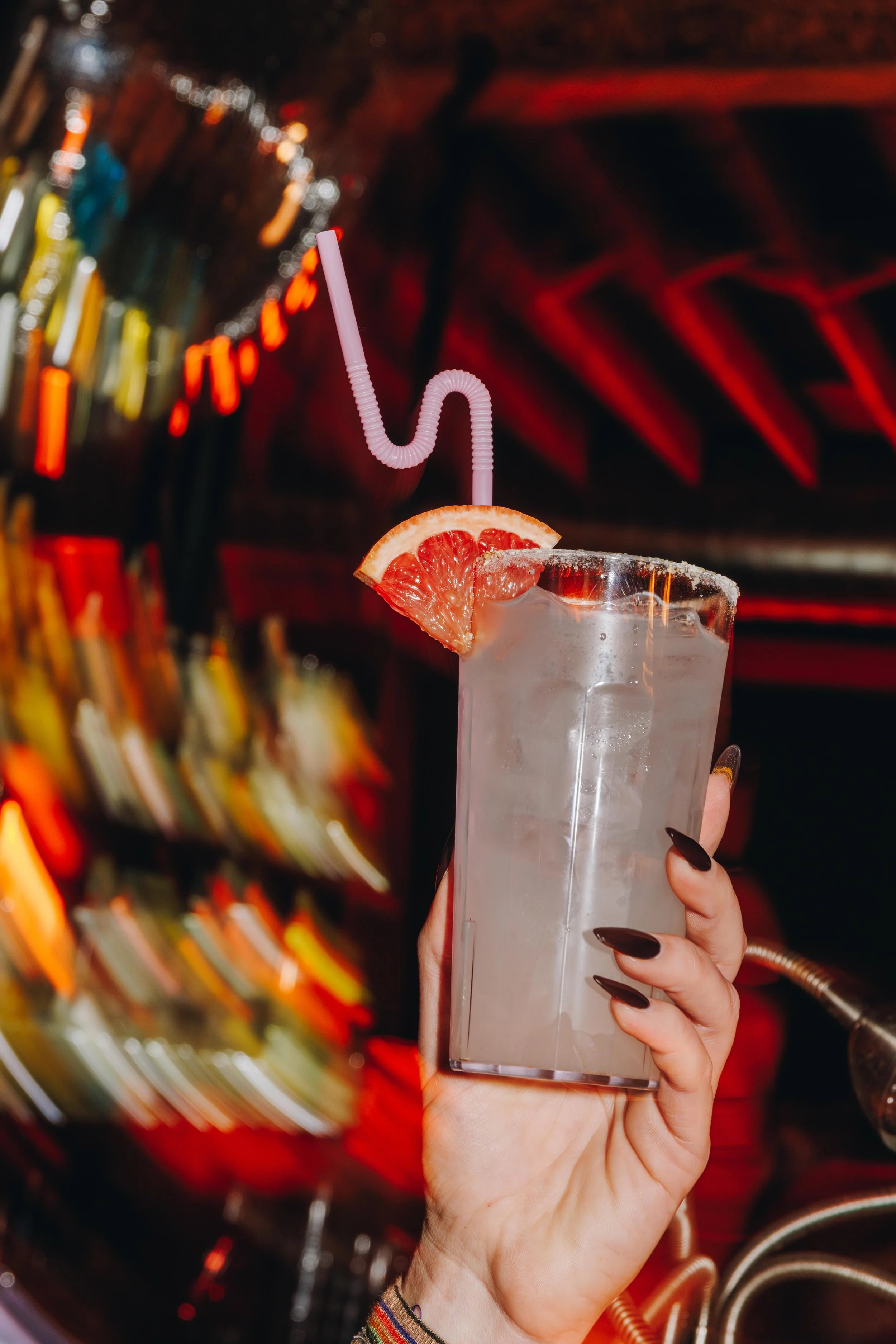 A person's hand with black nail polish holding a tall glass of a clear cold beverage garnished with a slice of grapefruit and a pink bendy straw, set against a colorful, blurred background with neon lights.
