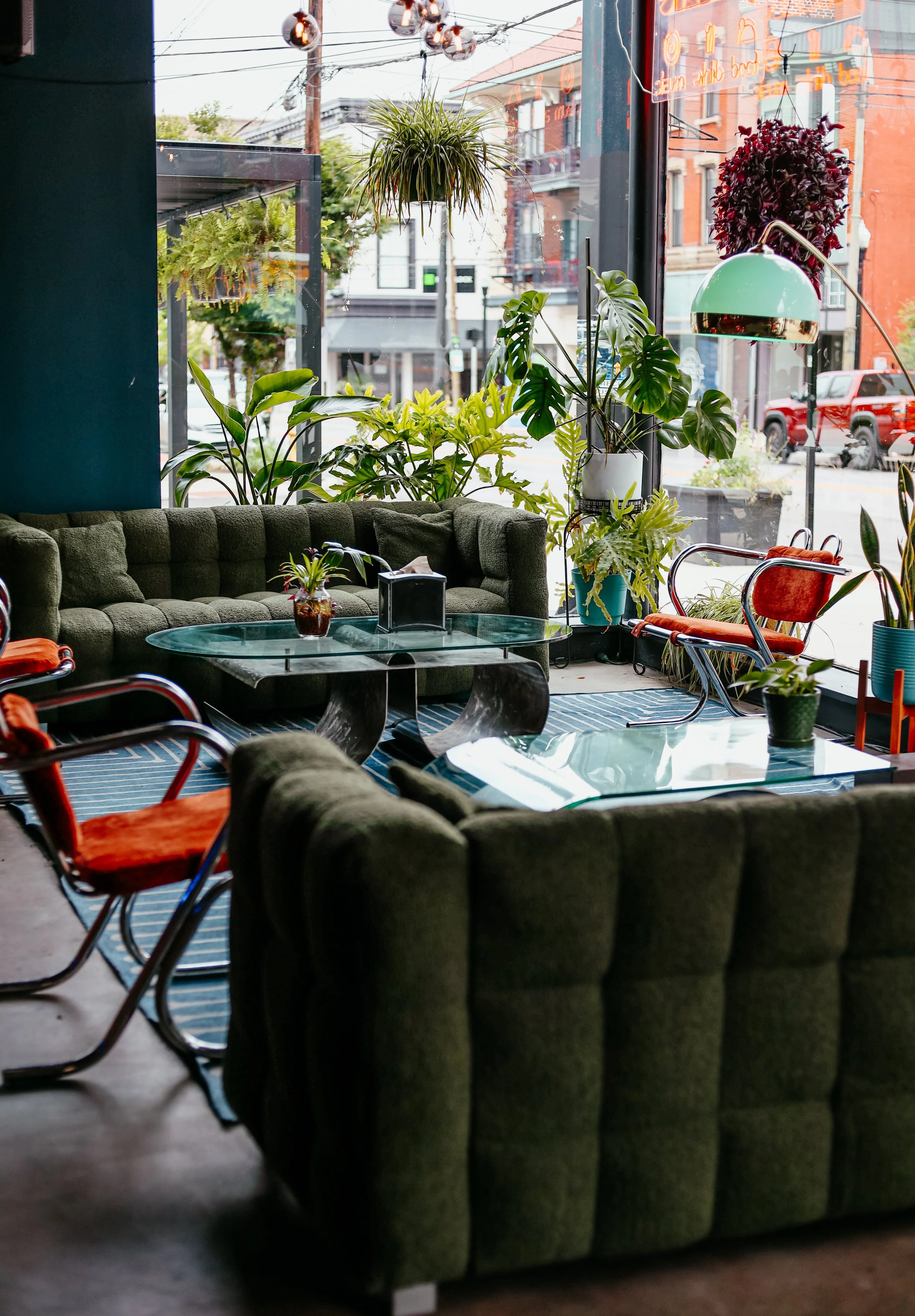 A cozy indoor seating area with a green velvet sofa, red chairs with metal frames, a glass table, and various green plants. Large window view of street outside with buildings, parked trucks, and power lines.