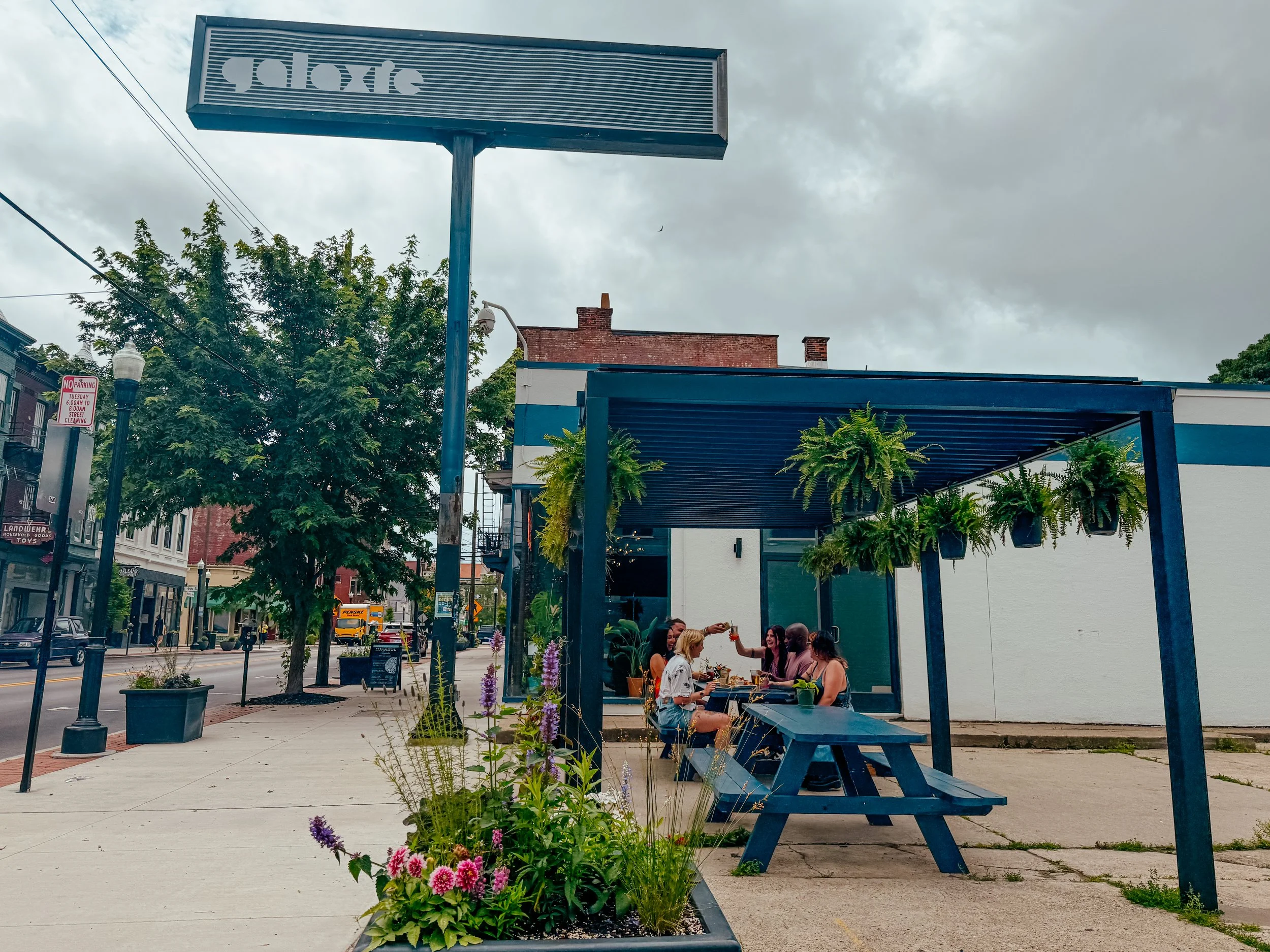 Group of people sitting at an outdoor table under a blue pergola with potted plants and flowers nearby, in an urban area with trees, buildings, and cloudy sky overhead.