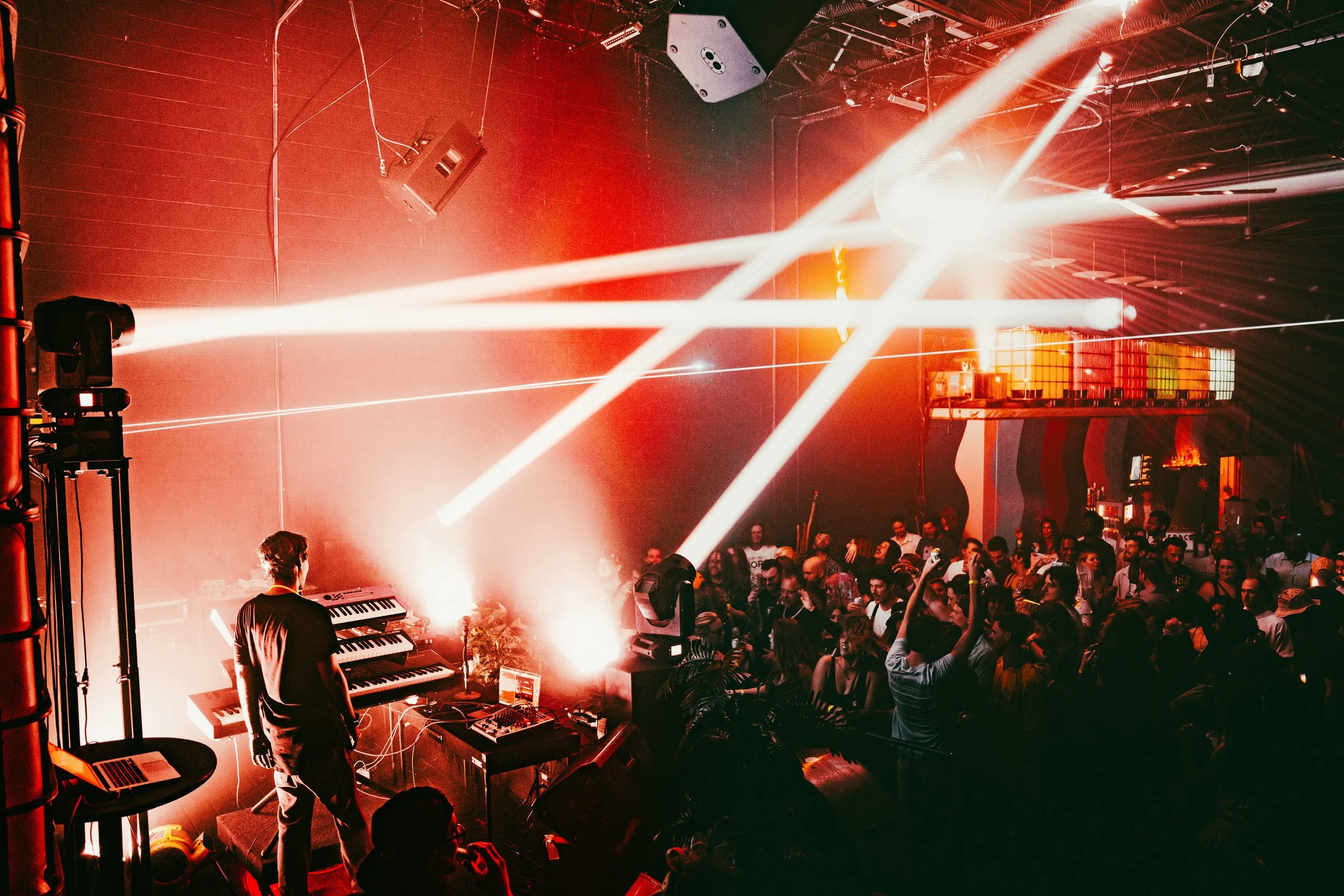 A nightclub scene with laser lights, a DJ playing electronic music, and a crowd of people dancing and enjoying the event. There are speakers, a keyboard, and lighting equipment visible.