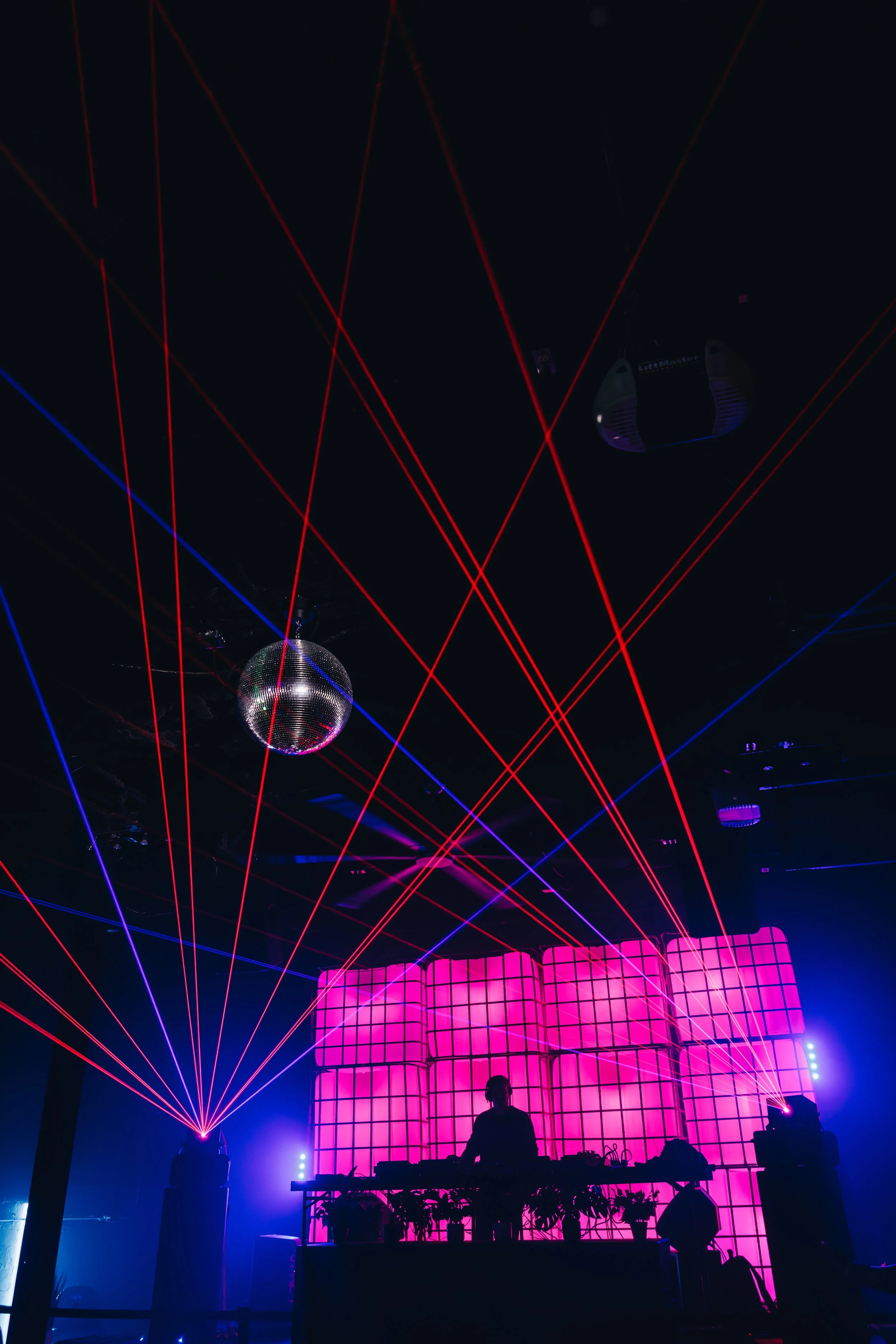 A DJ performs in a nightclub with red and purple laser lights, a disco ball, and a pink LED screen backdrop.